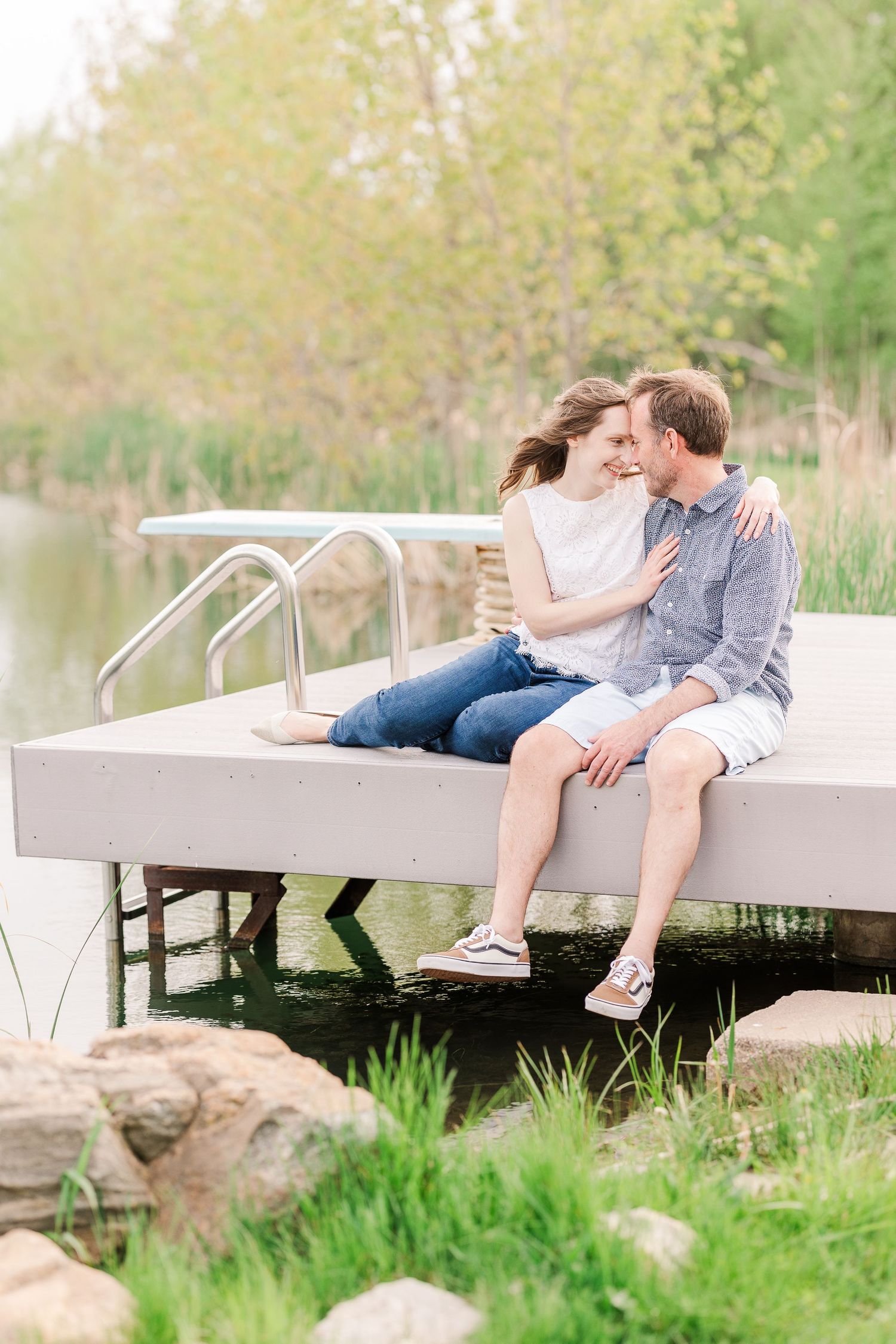A couple sits together on a dock by a peaceful pond surrounded by spring foliage.