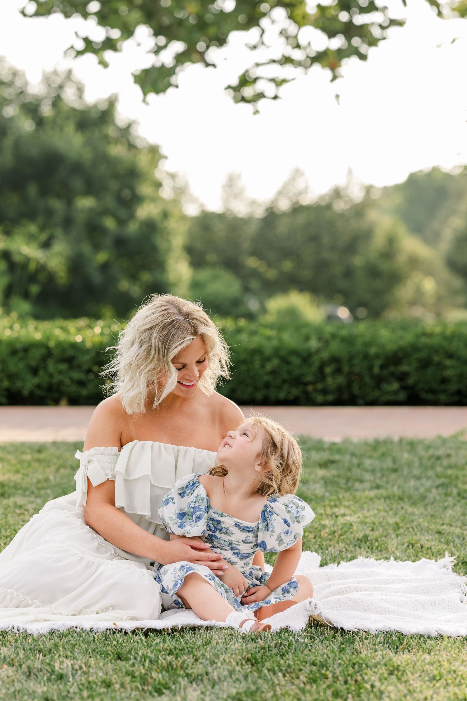 A smiling blonde mother in white off-shoulder dress sits on grass with her young daughter in a blue floral dress.