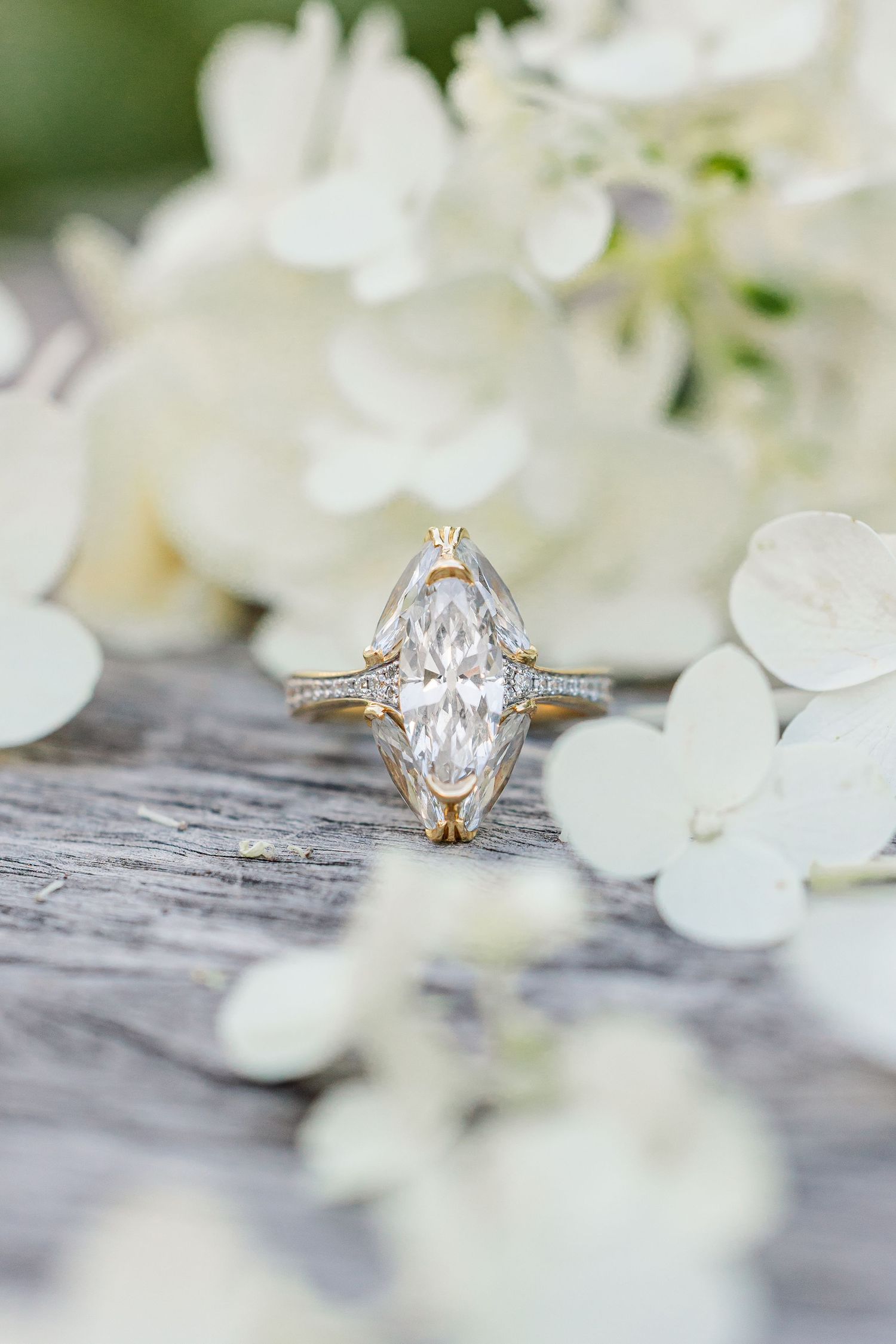 Elegant marquise diamond engagement ring in yellow gold setting displayed on rustic wood surrounded by white hydrangea flowers.