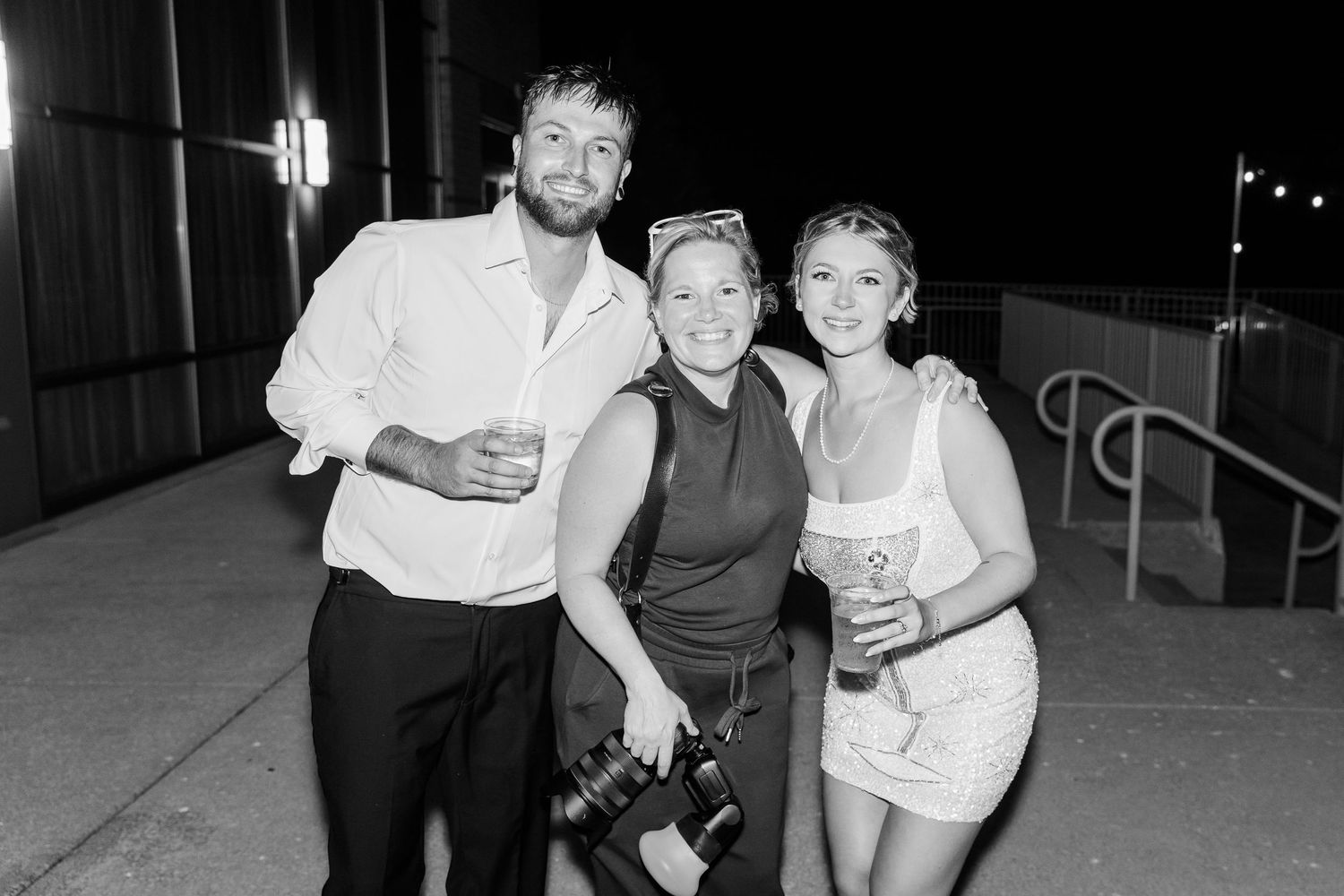 Black and white photo of three smiling guests posing together outdoors at evening wedding reception.