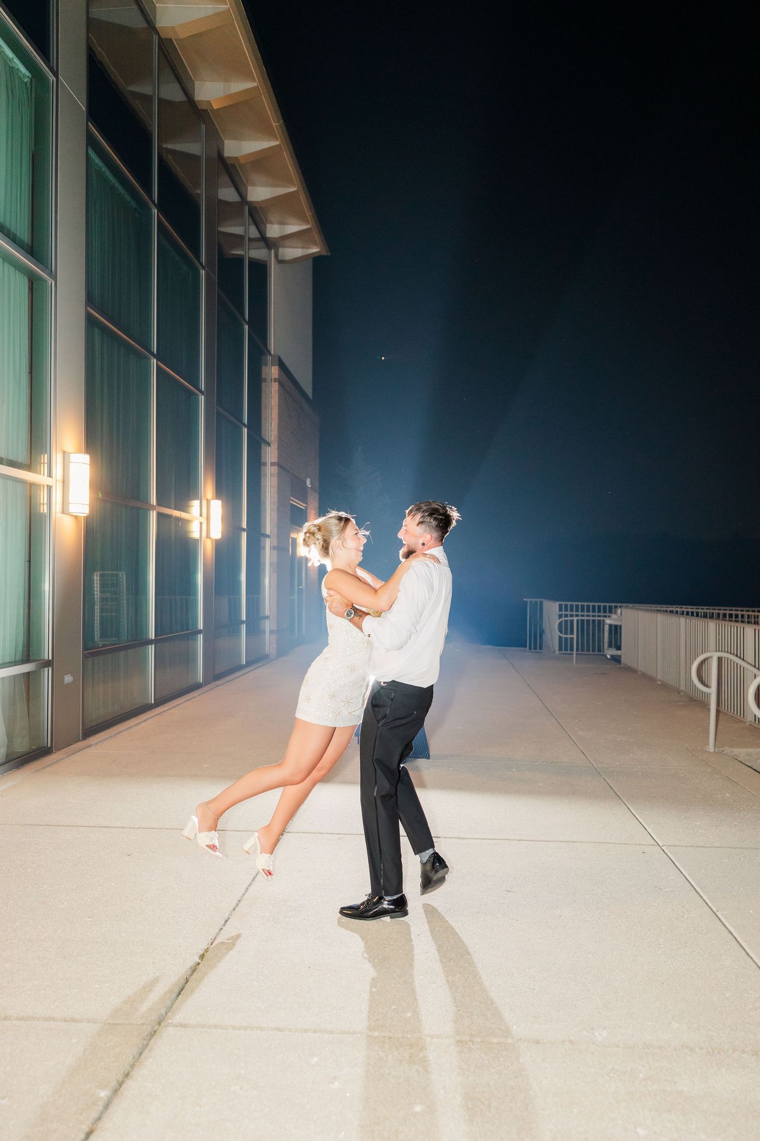 Couple dancing outdoors at night near a modern building, woman in white dress, man in formal attire, illuminated by soft lights.