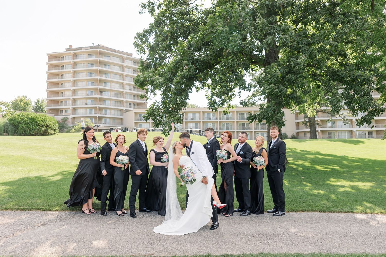 Wedding party in black attire poses with bride and groom on green lawn with hotel in background.