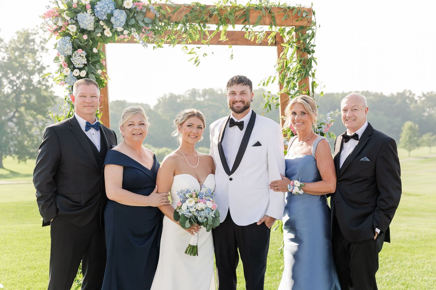 Wedding party family portrait of six people posing under a floral arch on a golf course.