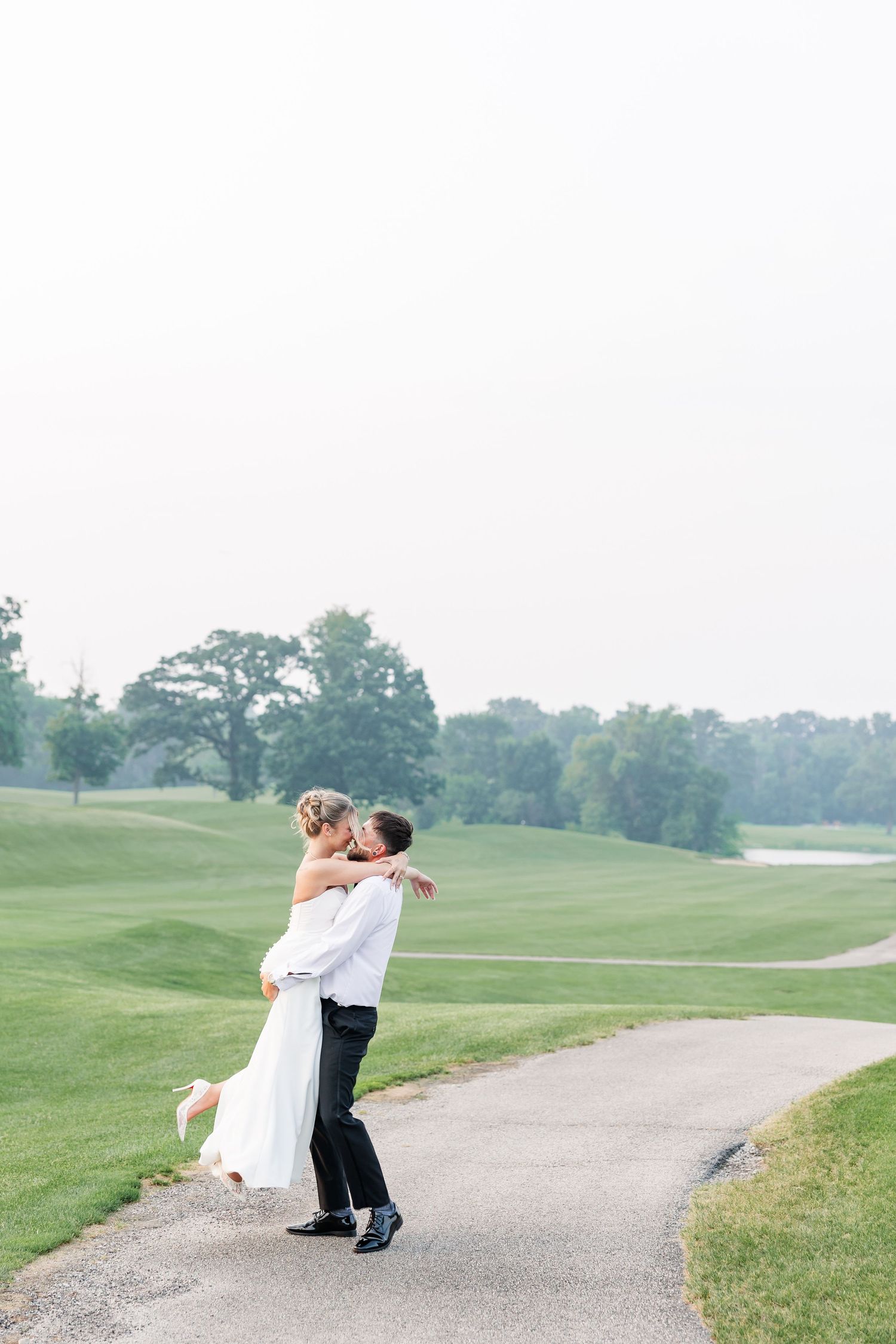 Groom lifting bride in white gown for a romantic kiss on a scenic golf course pathway.