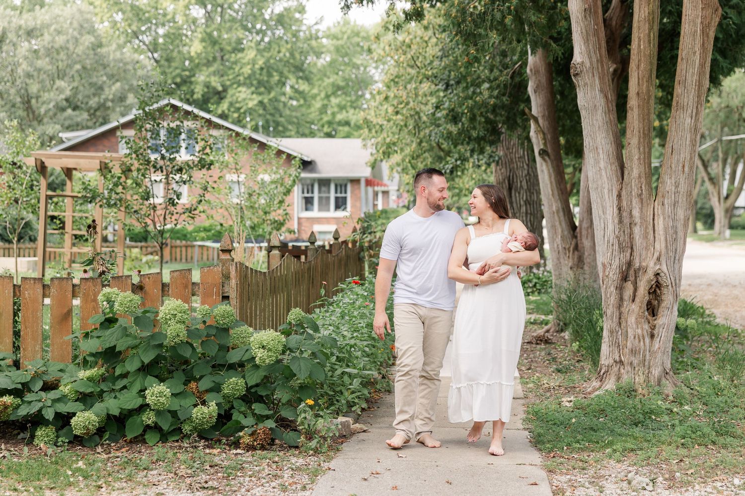 A couple walks along a garden path holding their newborn baby, smiling at each other near a wooden fence and lush greenery.