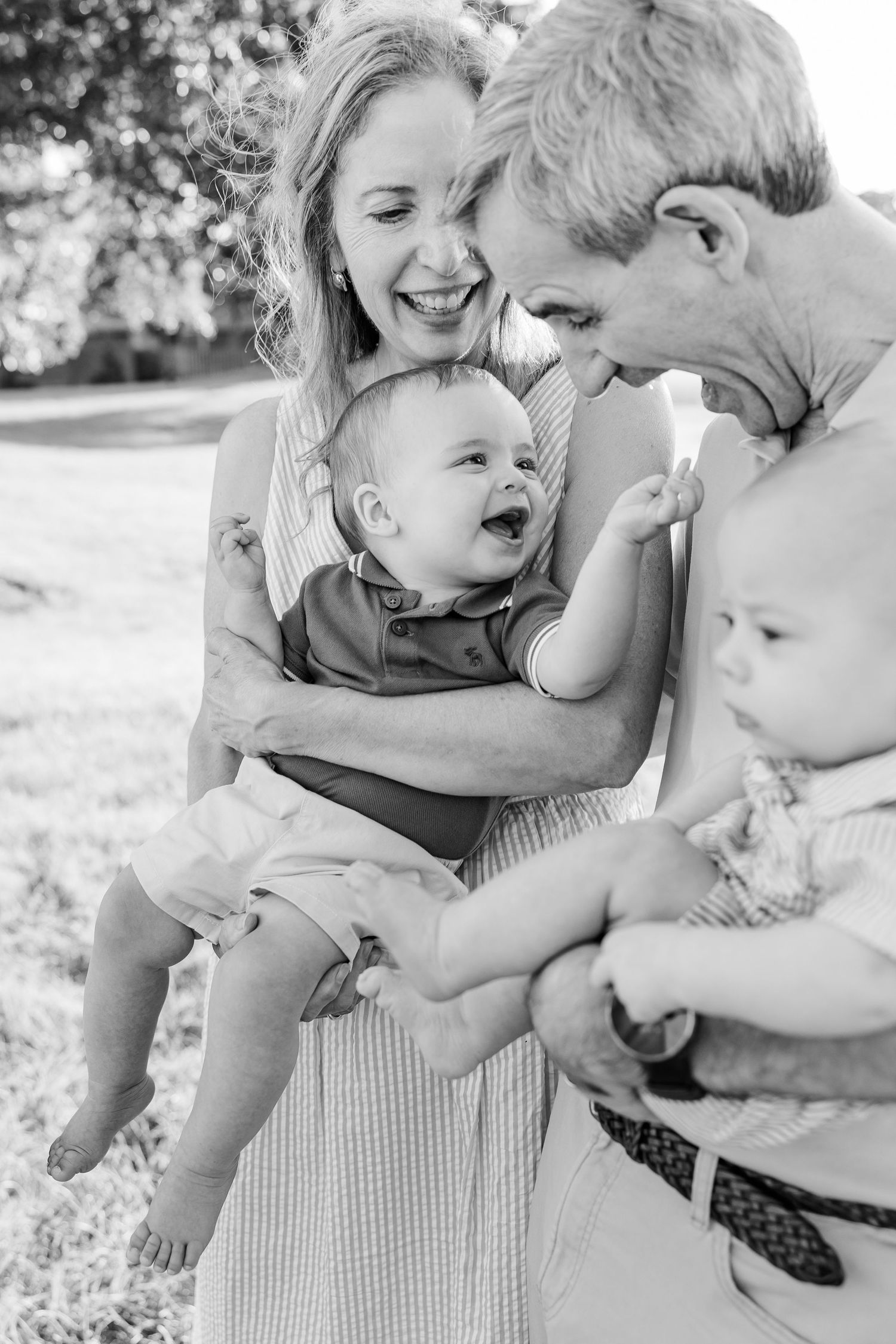 Black and white photo of grandparents laughing with twin babies outdoors in a sunny garden setting.