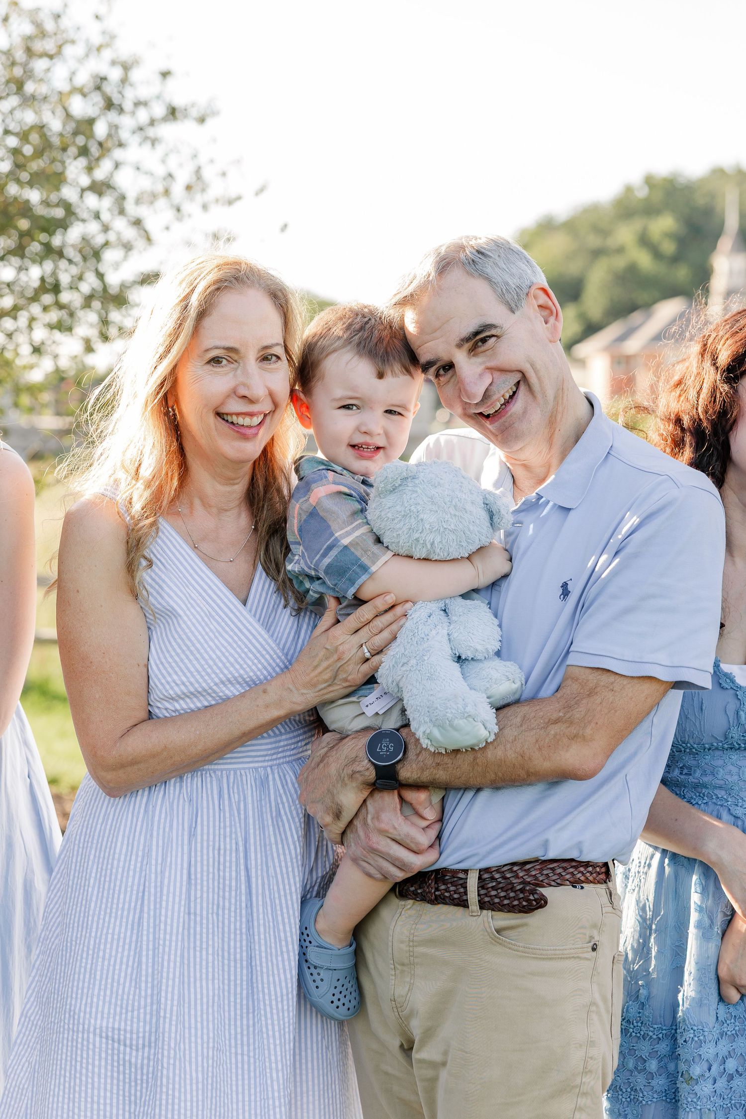 Smiling grandparents holding a happy toddler with a stuffed animal outdoors during a family photo session.
