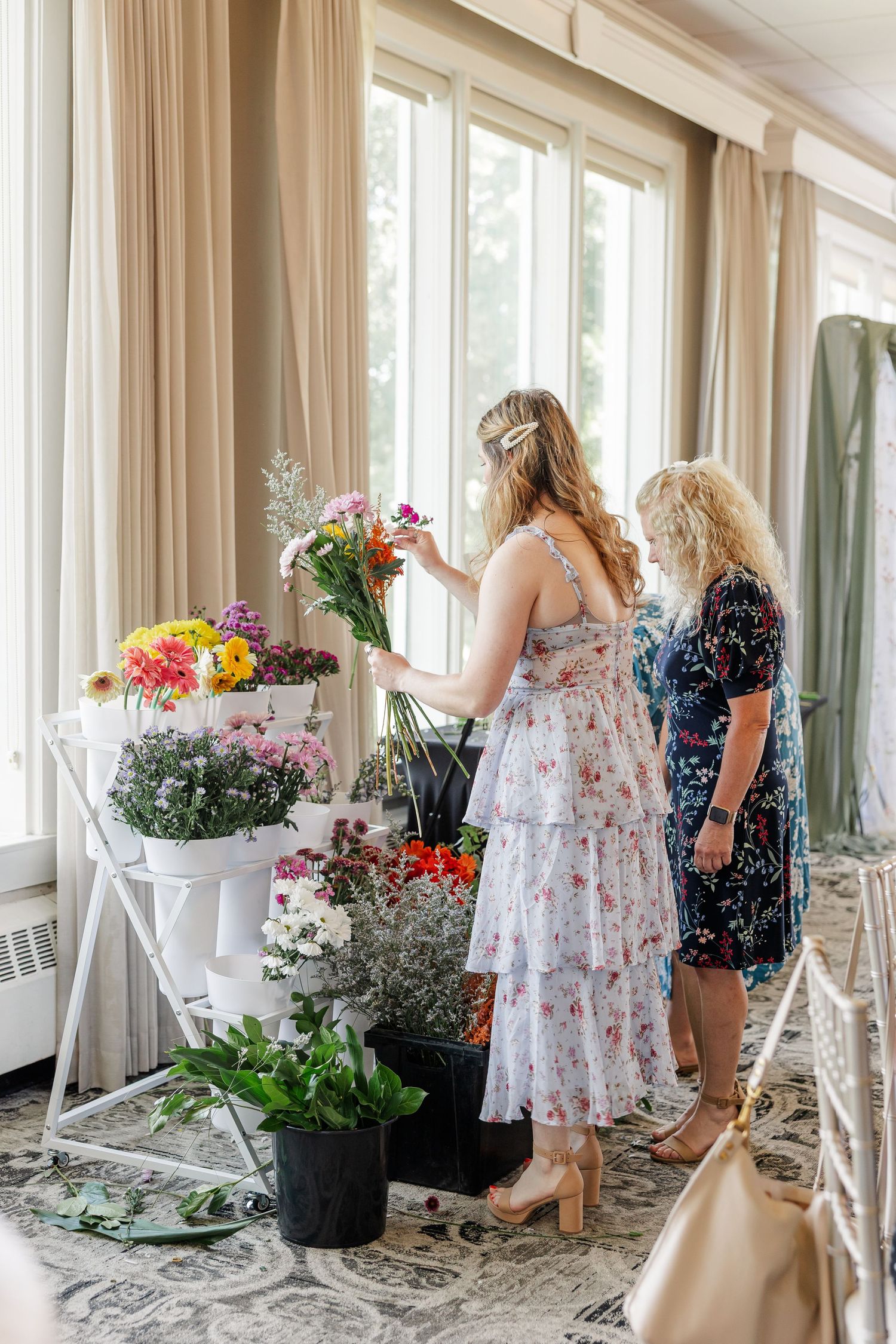 Two women selecting colorful flowers from a white display stand at an indoor floral arrangement activity.
