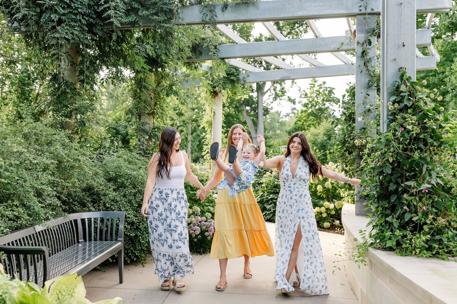 Three women in floral and yellow dresses laughing and dancing under a garden pergola together.