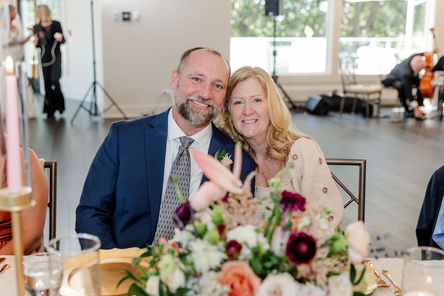 A smiling man in a navy blazer and a woman with blonde hair pose together behind a floral centerpiece at a wedding reception.