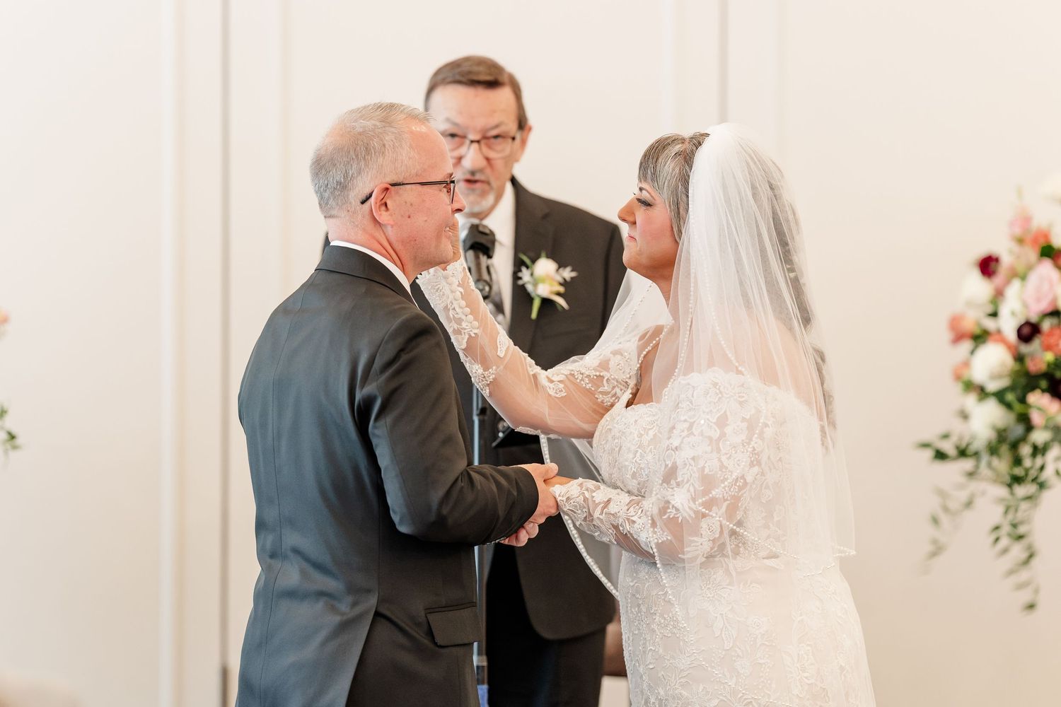 Bride in a lace gown and veil holds hands with the groom as an officiant looks on during their wedding ceremony near floral arrangements.