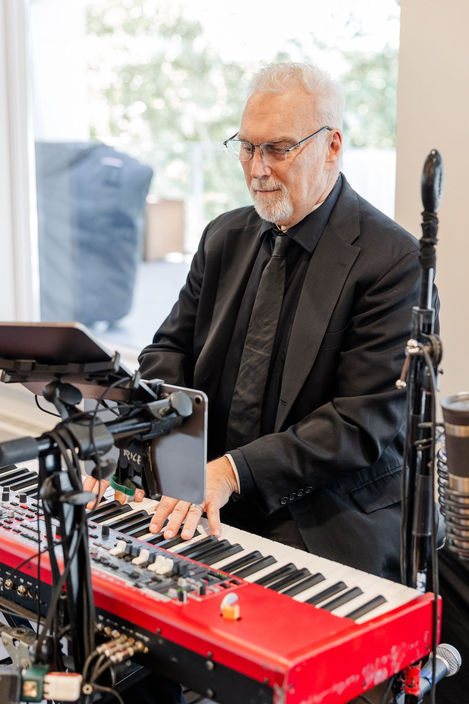 Gray-haired man with glasses and a goatee, dressed in a black suit, plays a red electric keyboard near a microphone stand at an event.