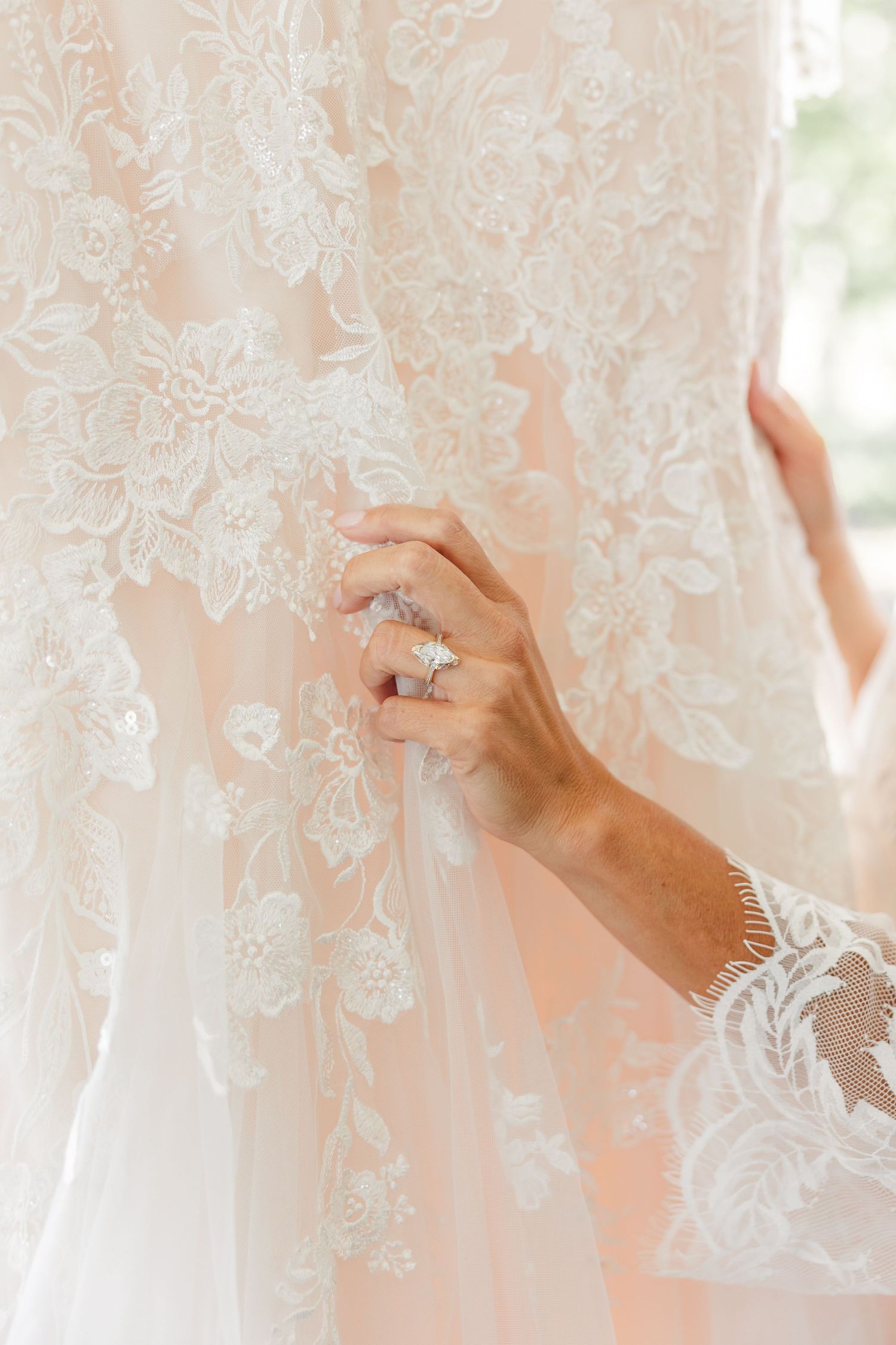 Close-up of a woman's hand with a diamond ring touching the delicate lace appliqué on a blush wedding gown.