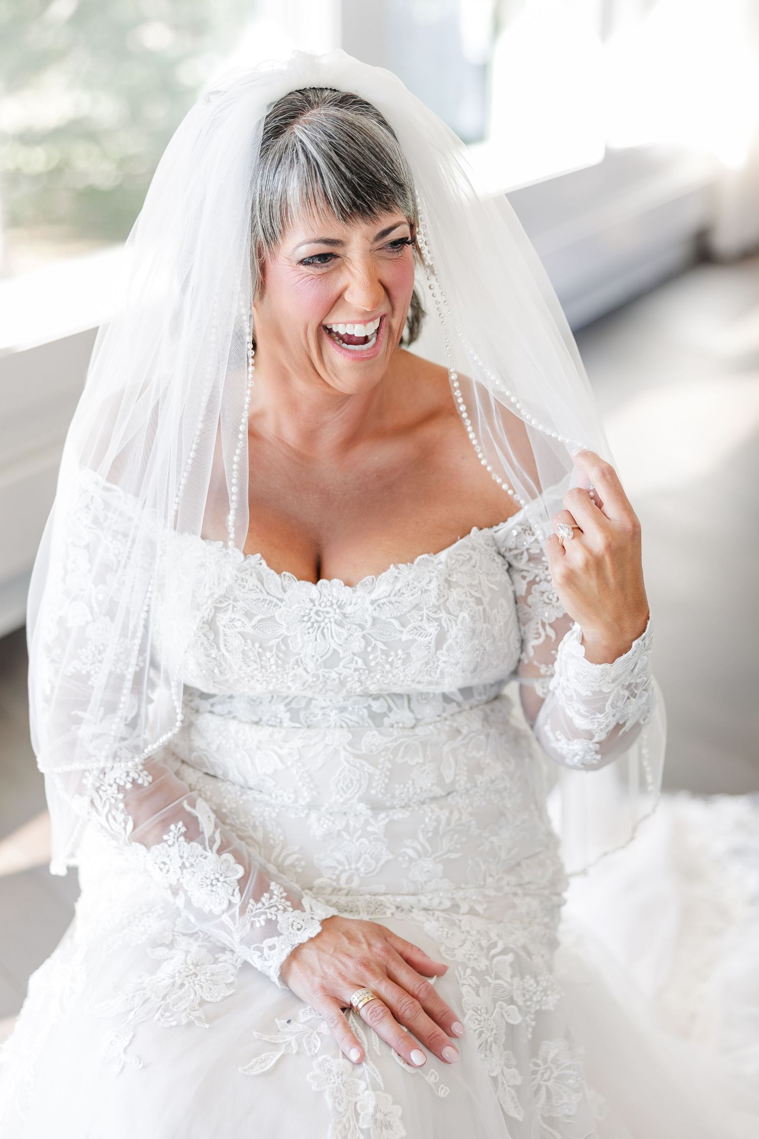 Smiling bride in an off-the-shoulder lace wedding gown and long veil laughs joyfully while seated indoors.