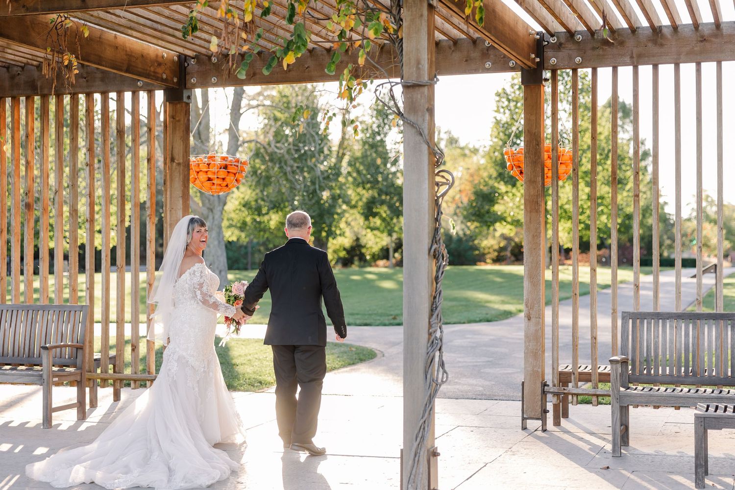 Bride in a white lace gown holds hands with a groom in a dark suit as they walk through a wooden pergola toward a sunlit garden.