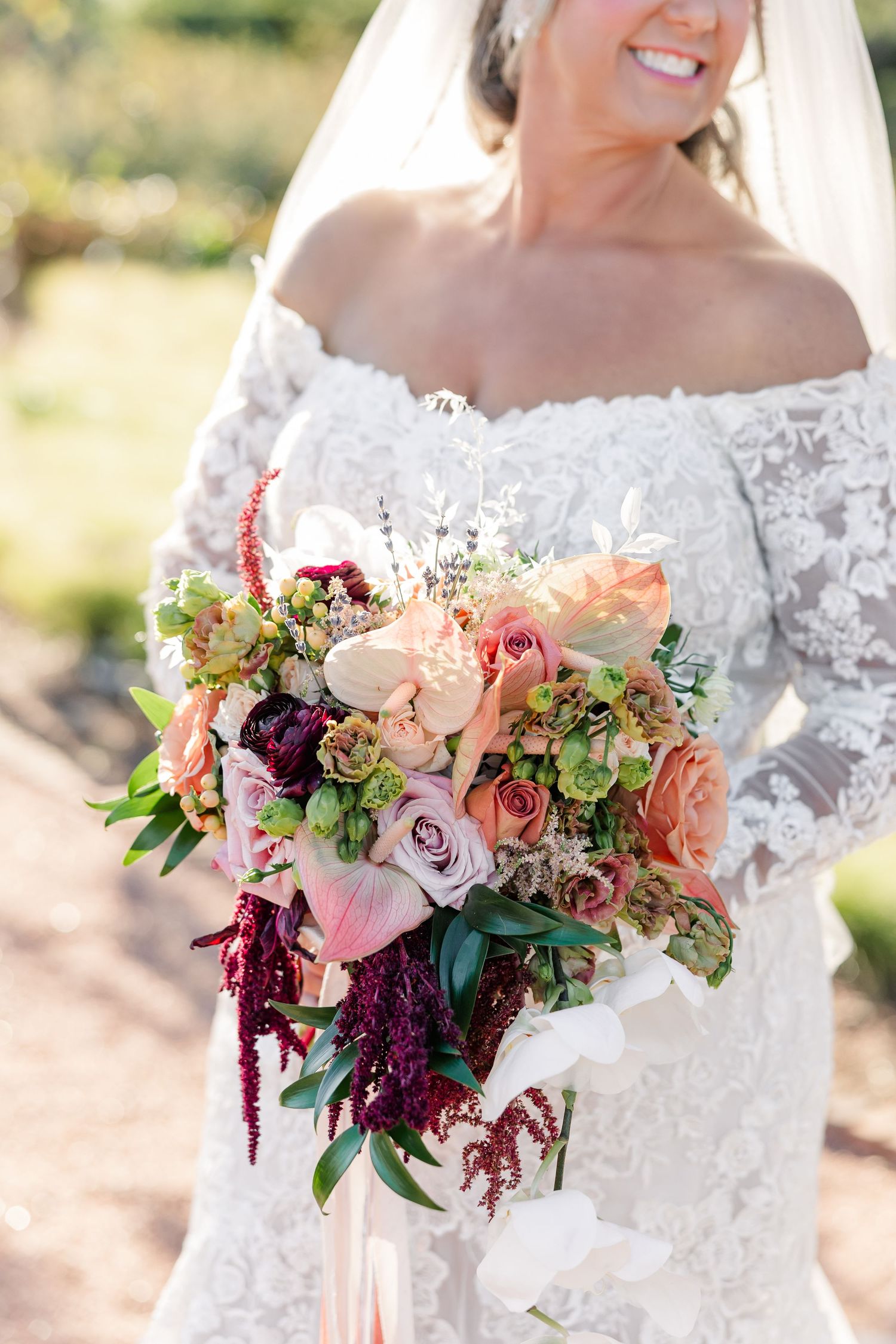 Smiling bride in an off-the-shoulder lace wedding gown holds a lush bouquet of blush roses, succulents, and burgundy amaranthus.