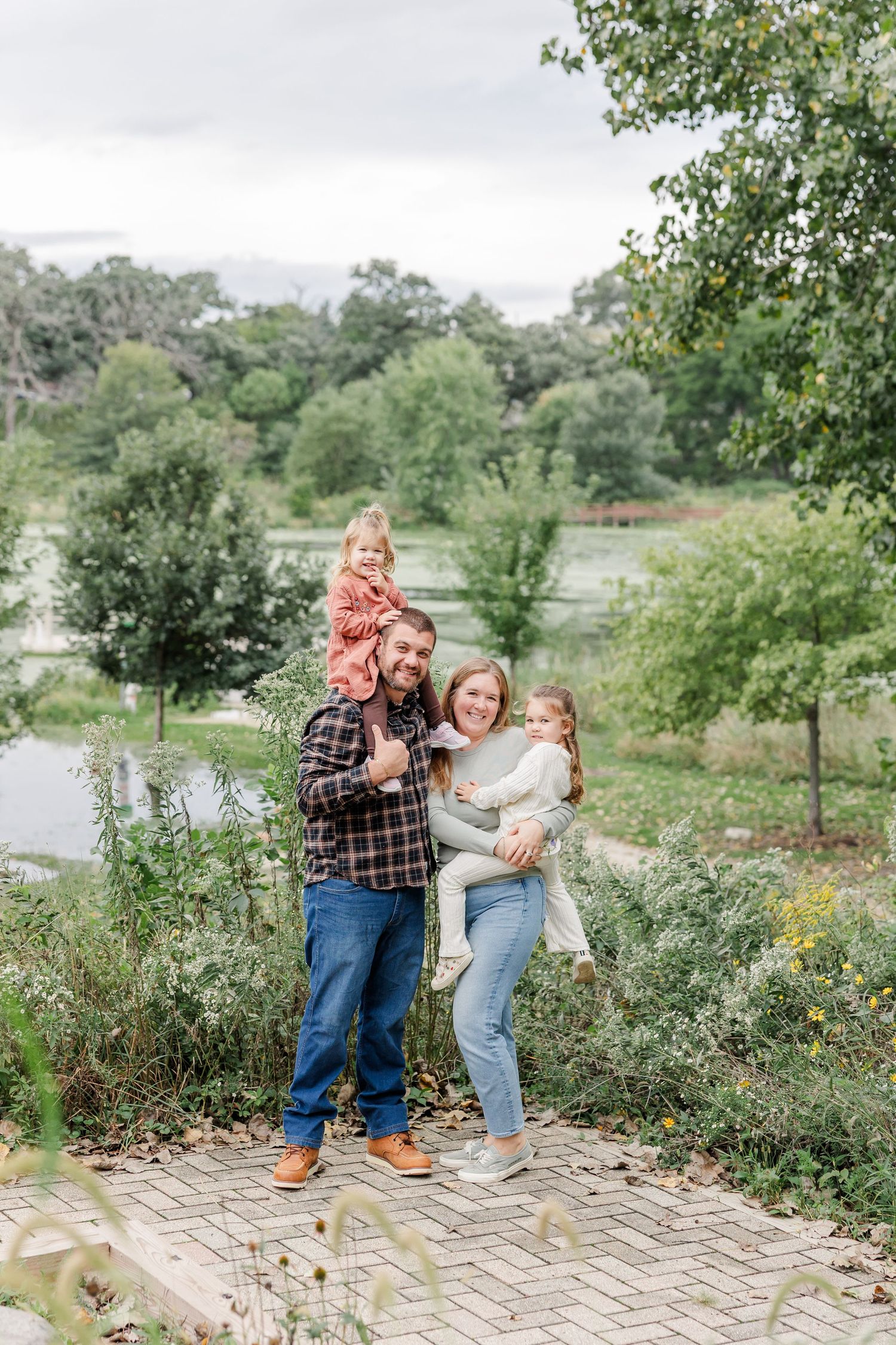 Happy family of four posing outdoors near a pond, dad with toddler on shoulders, mom holding young child, lush greenery background.