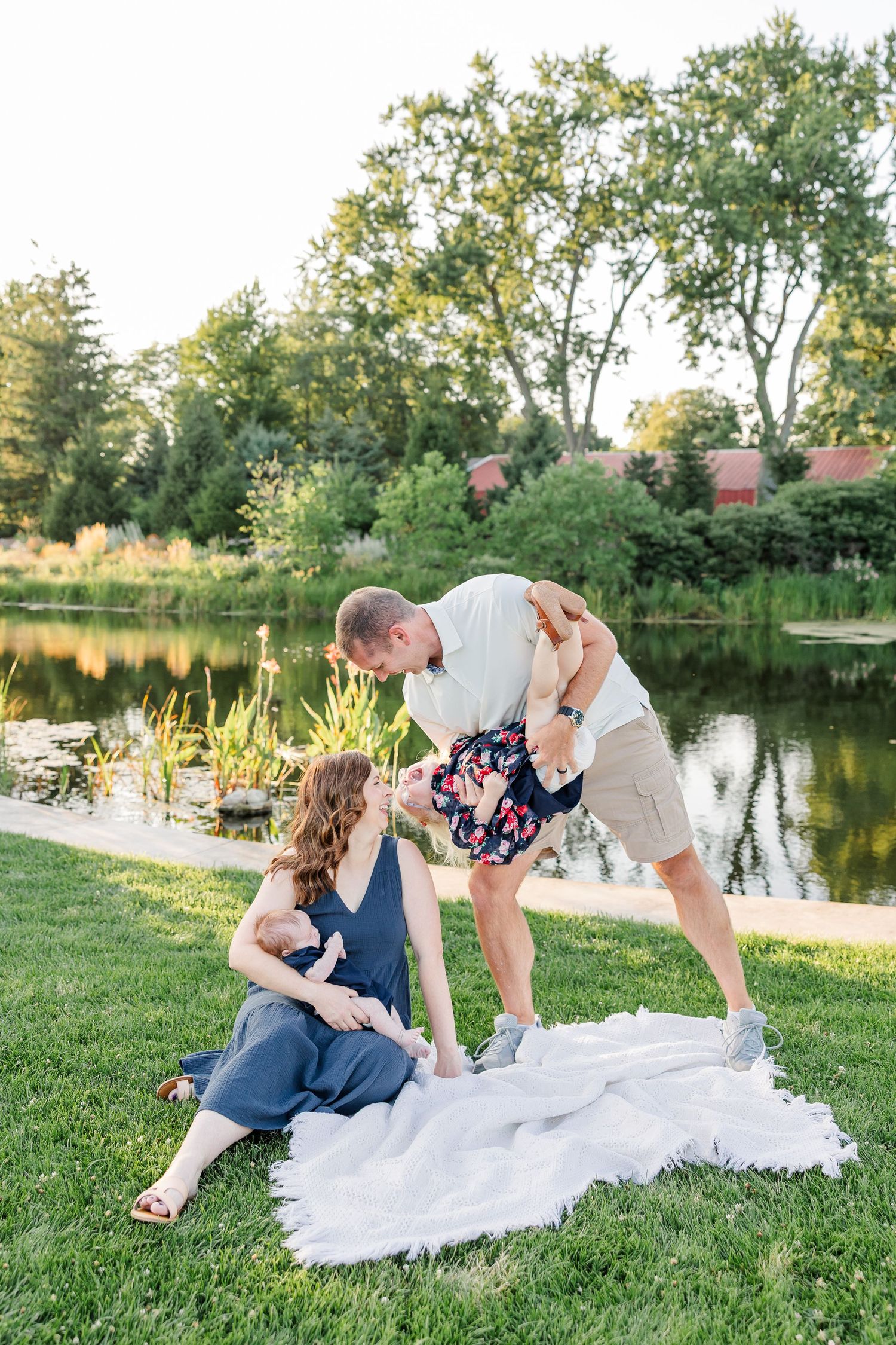 Andrew playing with Caroline as Hilary sits with newborn Lincoln on a blanket beside the pond.