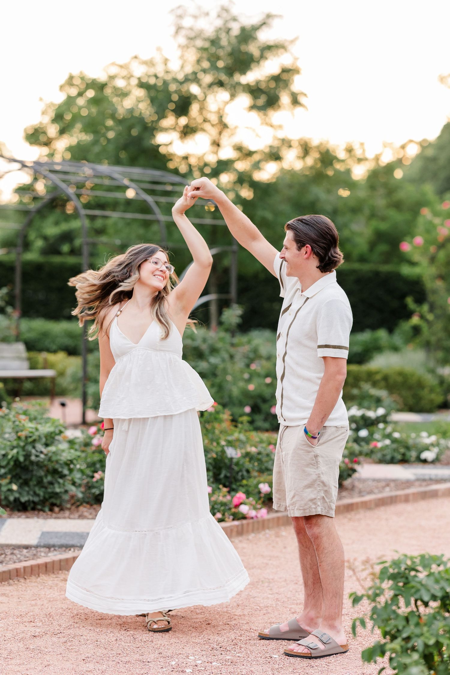 A couple dances joyfully in a garden at sunset, the woman in a white dress being twirled by a man in casual attire.