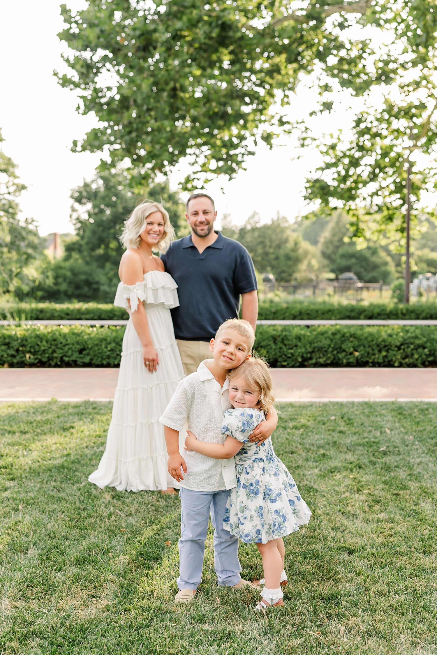A joyful family of four poses outdoors, with two young children hugging while parents stand behind them on green grass.