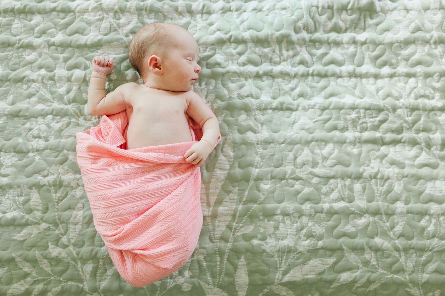 A peaceful newborn baby partially wrapped in a pink swaddle rests on a green floral quilted blanket.