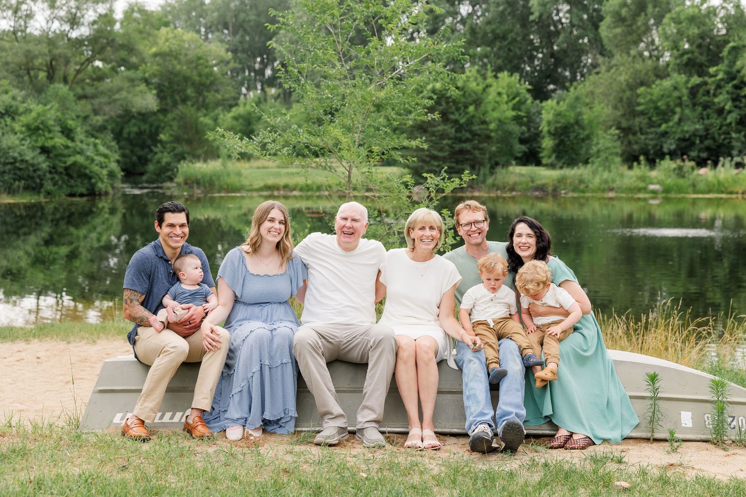 Extended family group portrait of eight people seated on bench near peaceful pond with green trees.