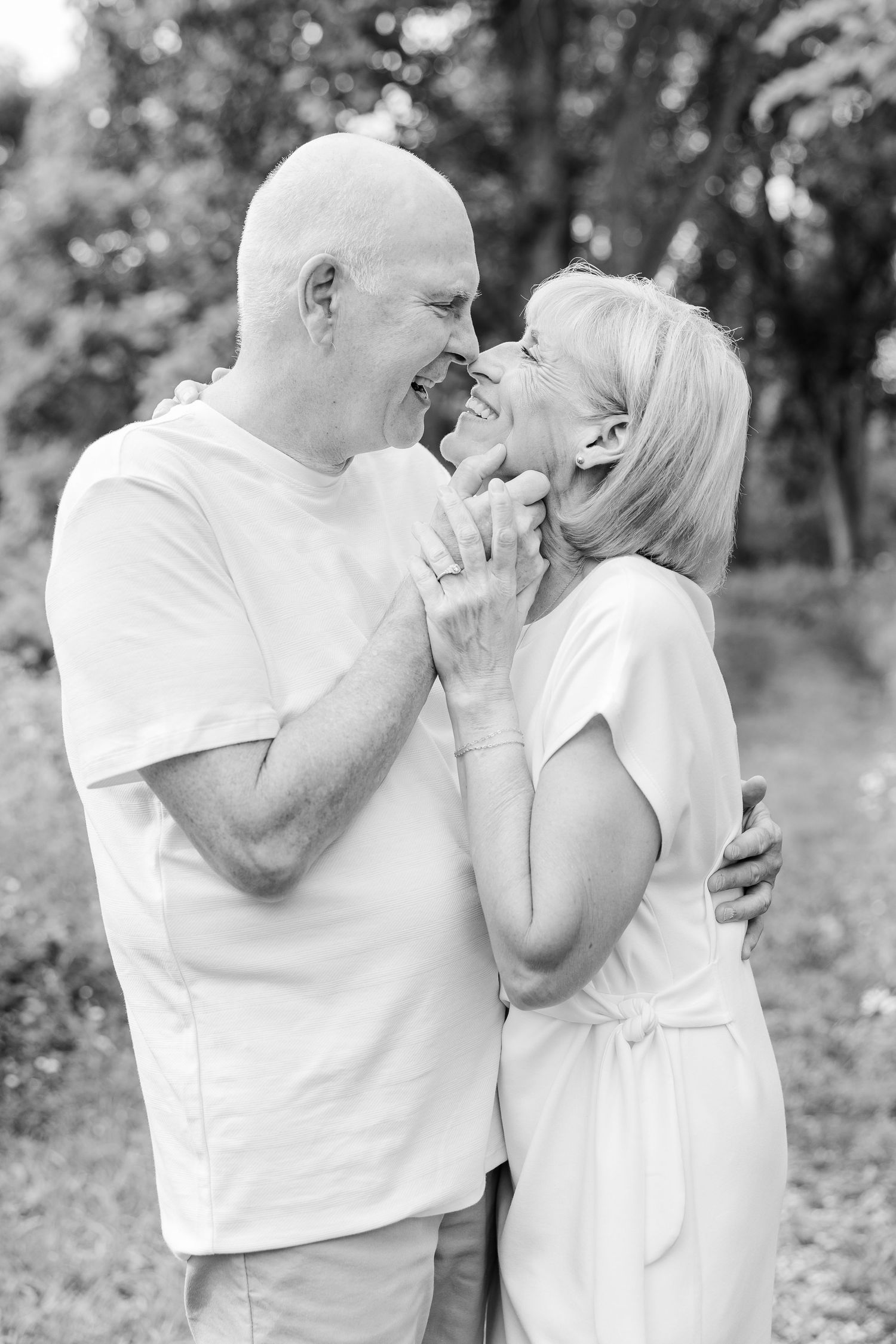 Black and white photo of a happy older couple laughing and embracing outdoors in a lush garden setting.