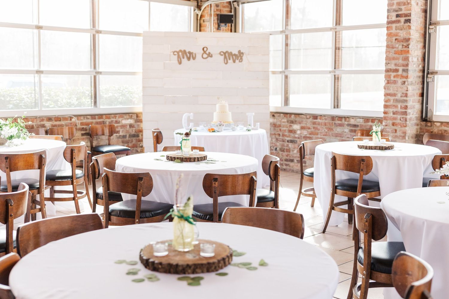 Rustic wedding reception venue with white tablecloths and wooden chairs in brick-walled room.