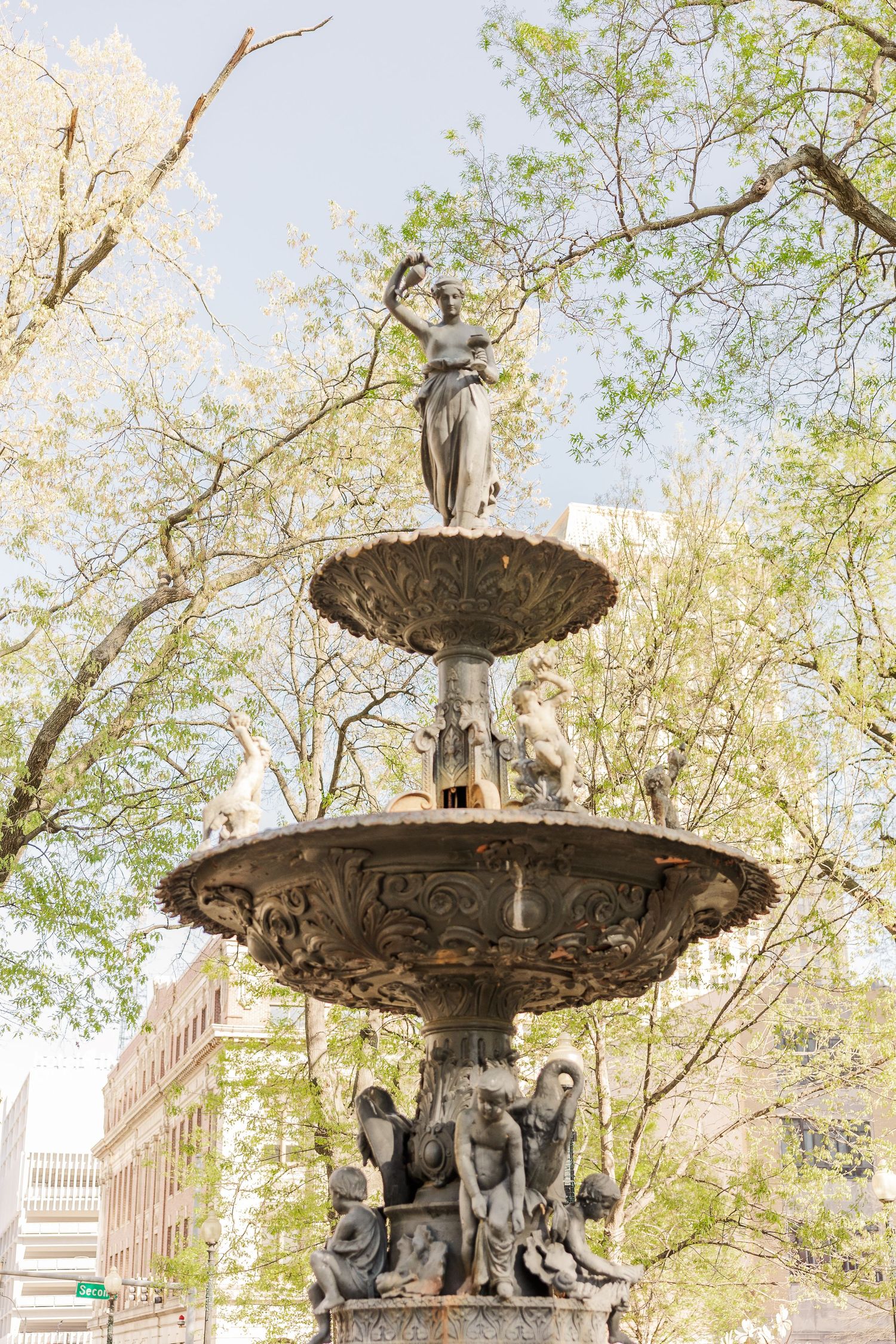 An ornate tiered fountain with classical statues stands among spring trees.
