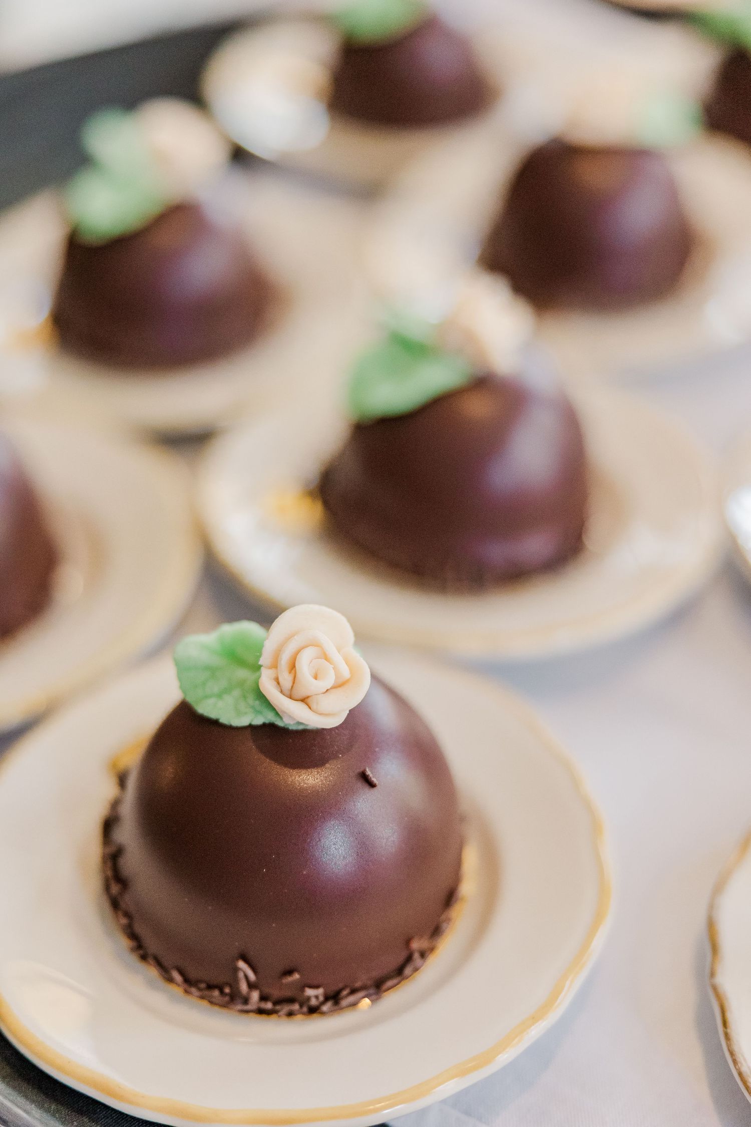 Chocolate dome desserts garnished with small sugar roses and mint leaves on cream-colored plates.