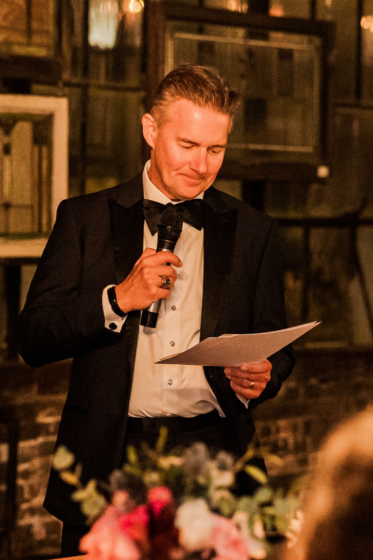 A speaker in a tuxedo delivers a speech while holding papers at an evening event.