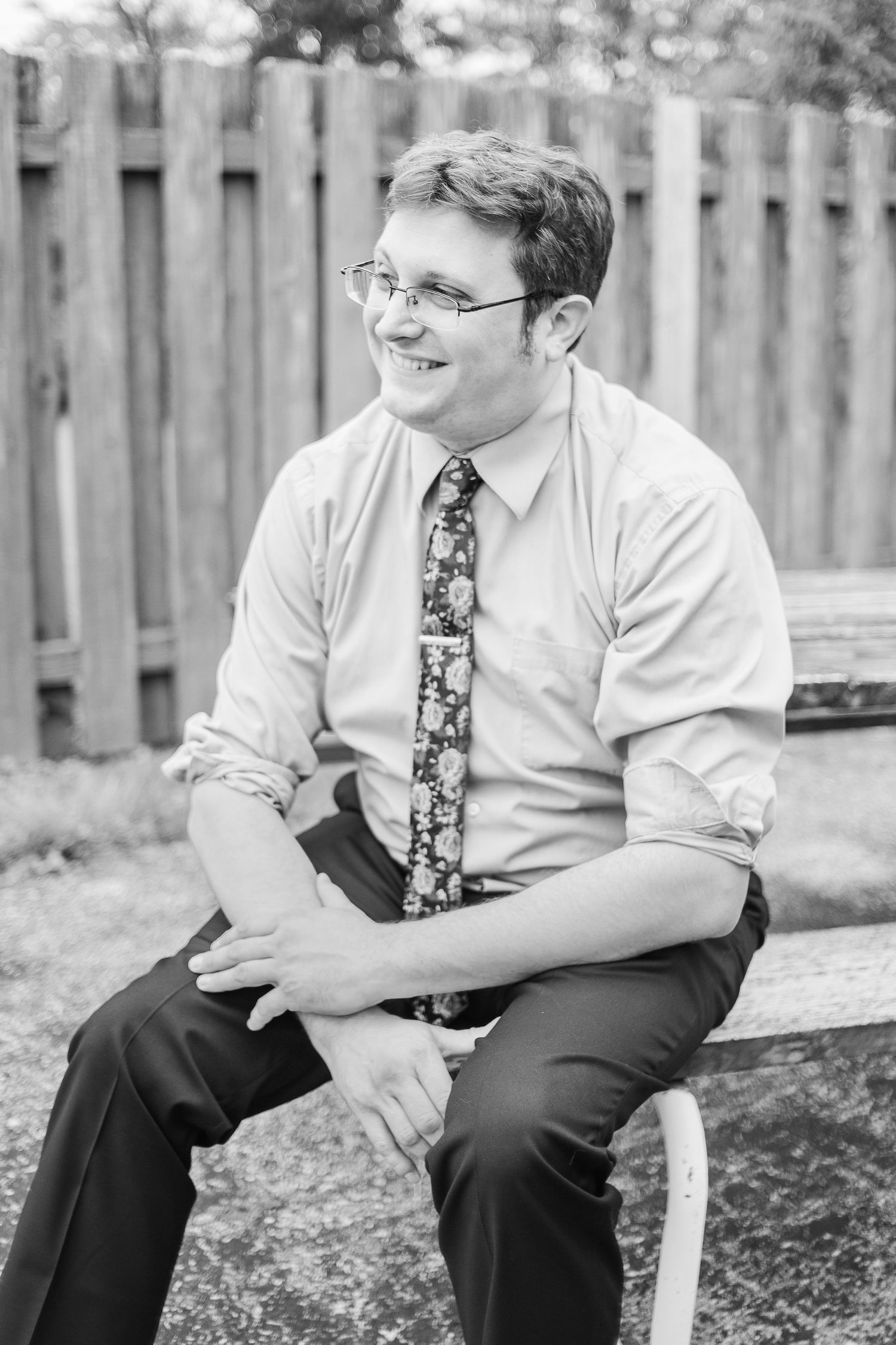 Black and white portrait of a person in business attire sitting outdoors by a wooden fence.