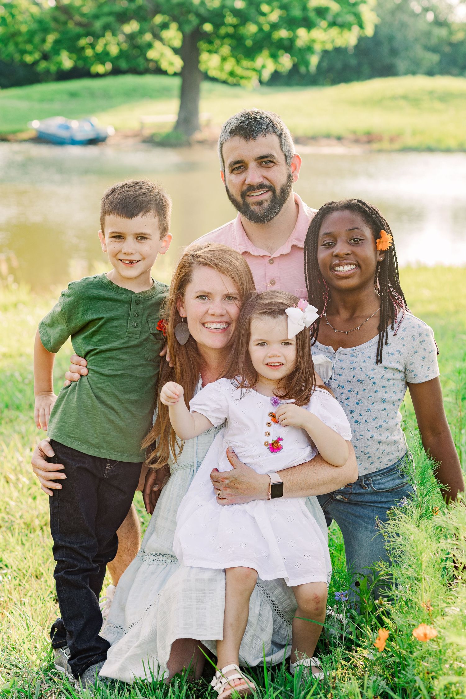 Happy family group poses together on grassy hill with pond in background.