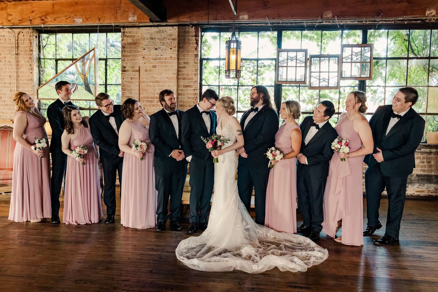 Wedding party stands in line against exposed brick wall and large windows in rustic venue.