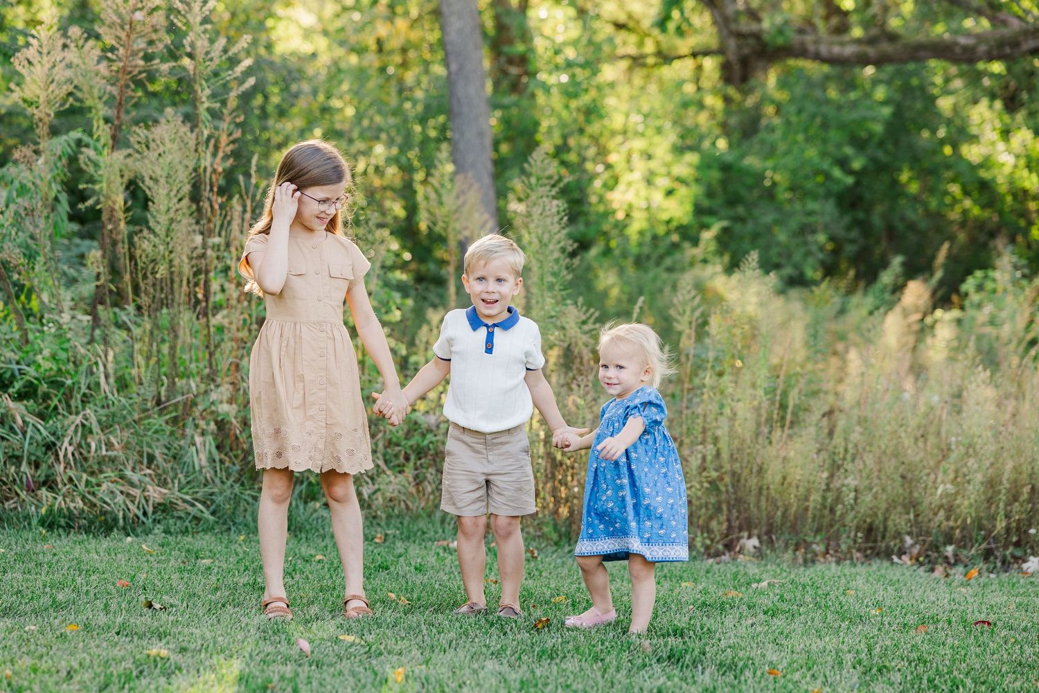 Three children stand together in a grassy field during golden hour lighting.