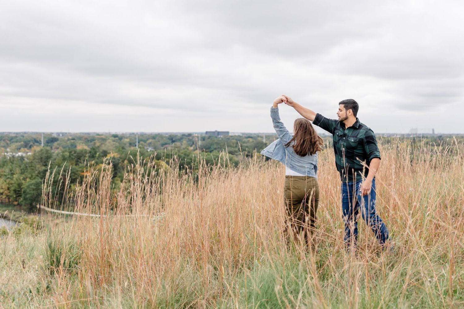 Couple dancing in a tall golden grass field on a hilltop overlooking trees under a cloudy sky, she twirls under his raised hand.
