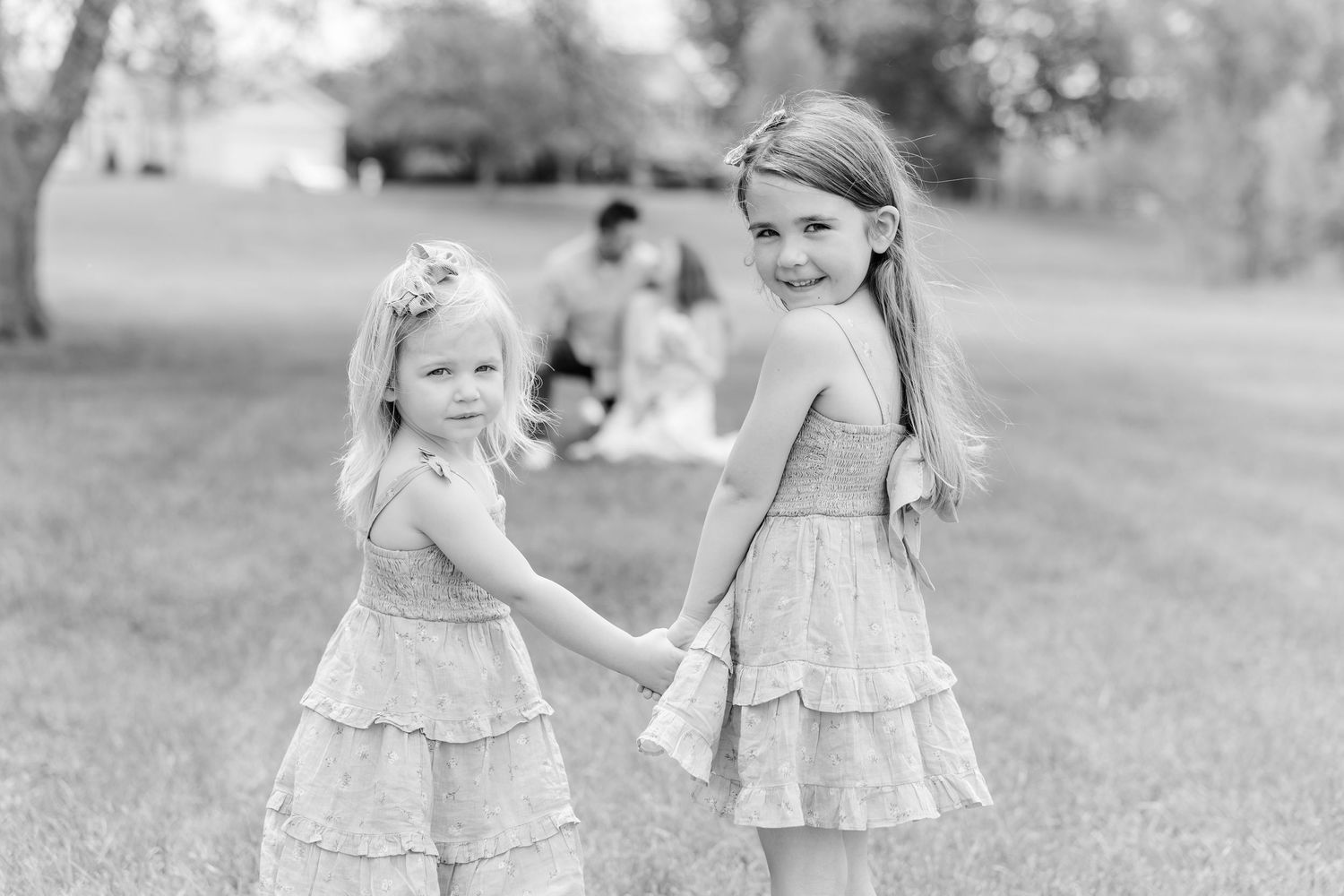 Two young girls in matching ruffled dresses hold hands in a park, smiling over their shoulders in a black and white photo.