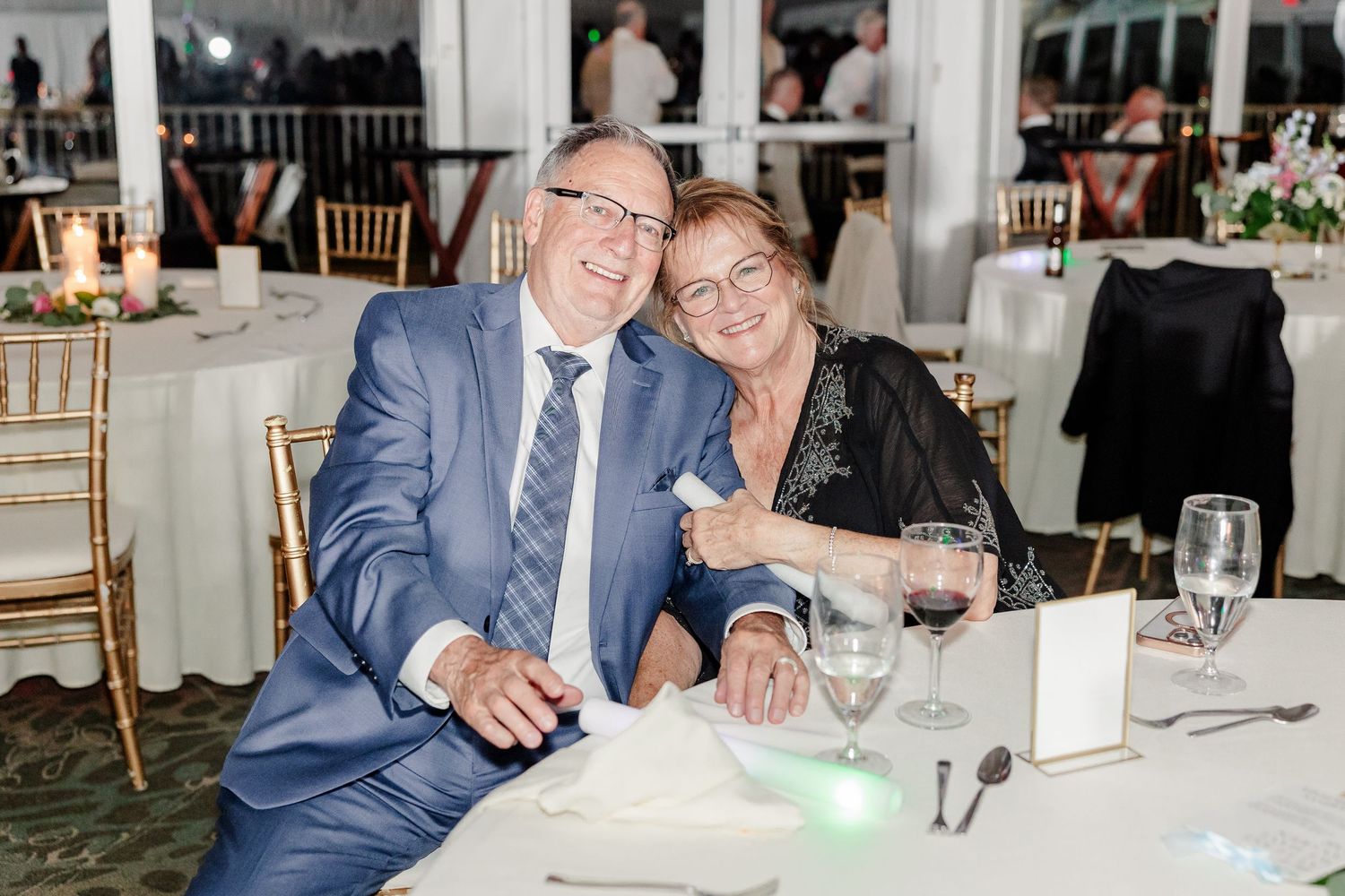 Smiling elderly couple in formal attire seated together at wedding reception table with wine glasses.