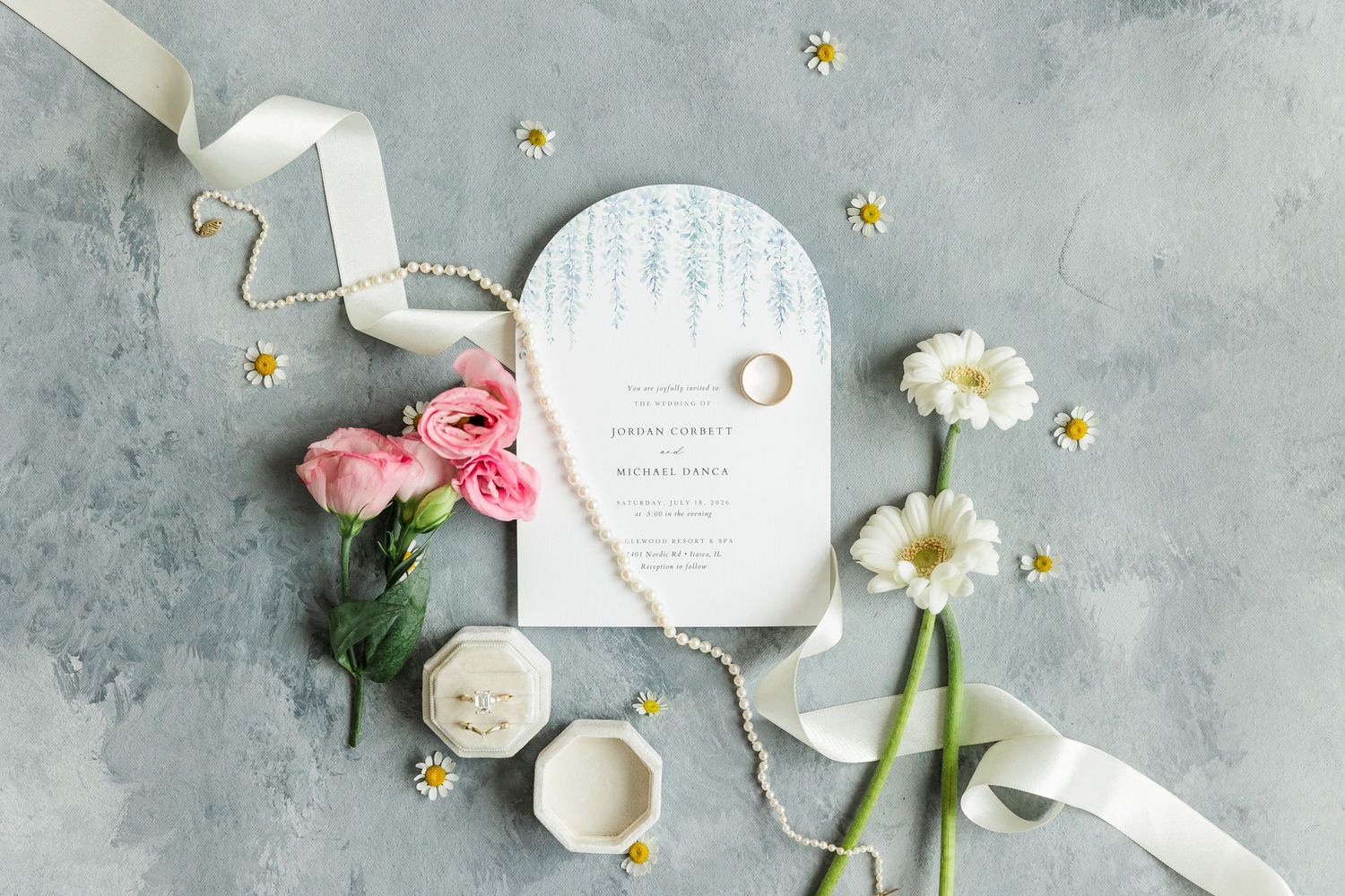 Elegant wedding flat lay with floral invitation, pink ranunculus, white daisies, pearl necklace, ribbon, and gold rings on gray background.