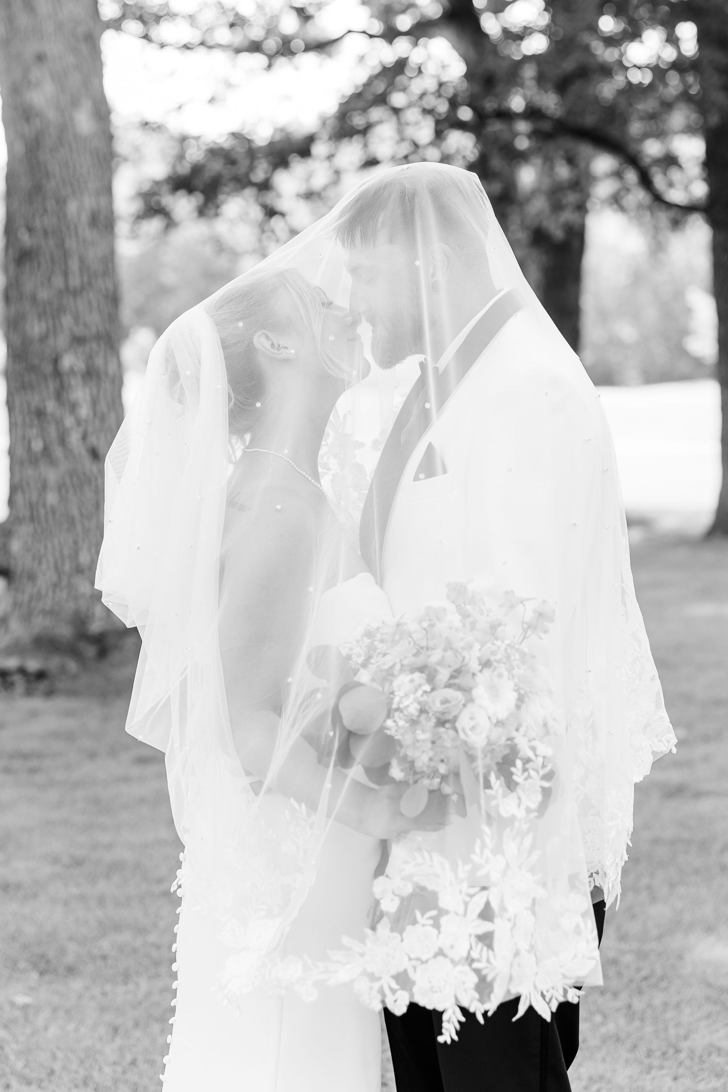 Bride and groom share intimate kiss under lace veil in black and white photo.
