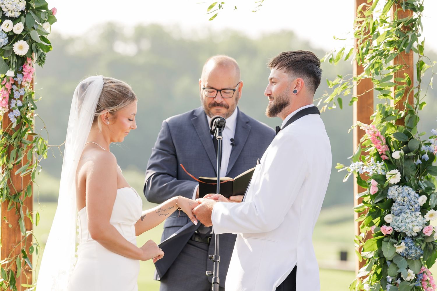 Bride and groom exchange vows outdoors at a floral arch with an officiant during a wedding ceremony.