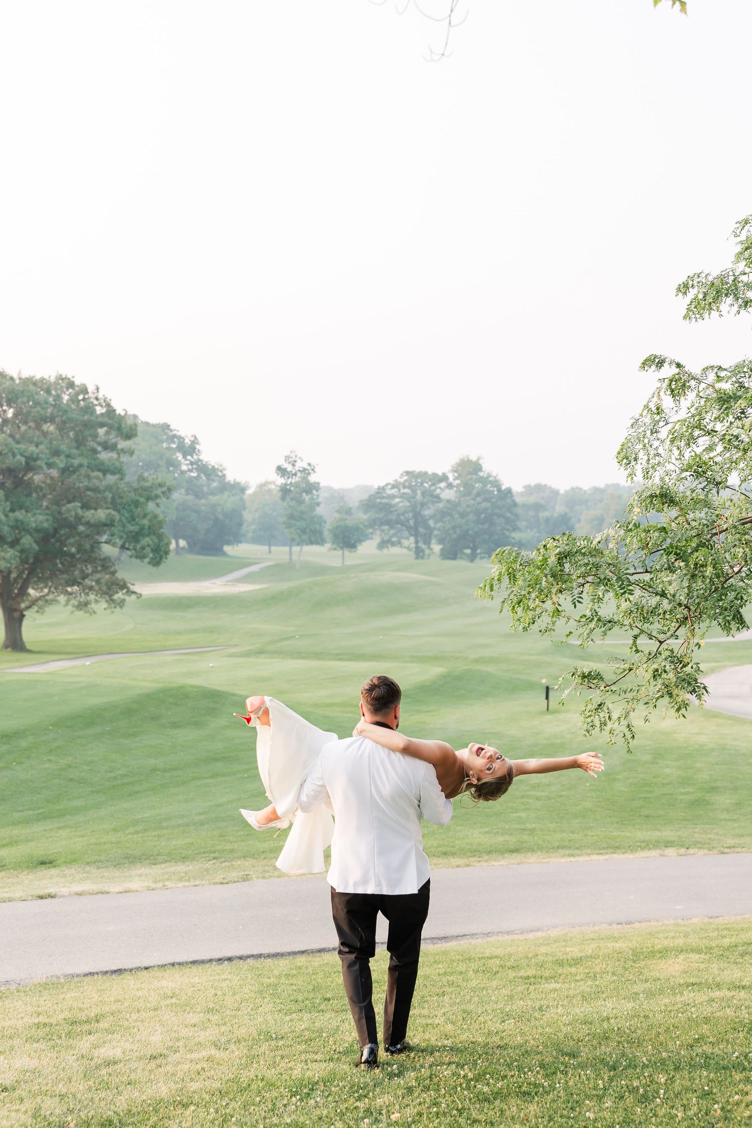 Groom lifting and spinning bride in white dress on lush green golf course wedding venue.