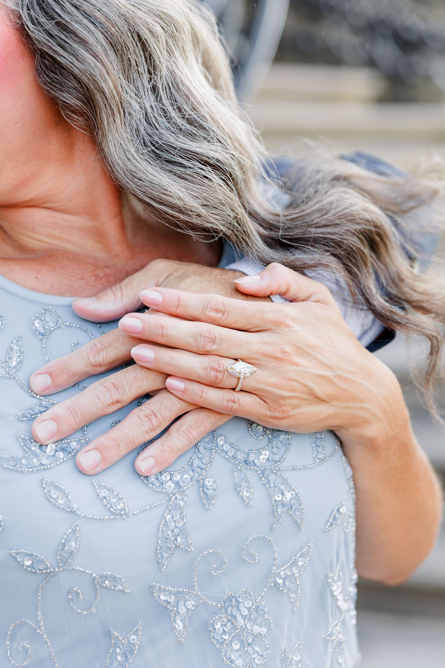 Close-up of a woman's hand with a diamond engagement ring resting on another person's shoulder in a blue beaded dress.