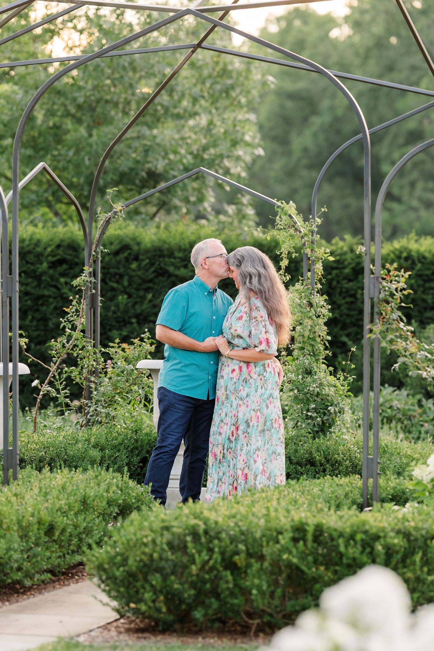 A romantic couple shares a kiss under a metal garden arbor surrounded by lush green hedges and climbing vines.