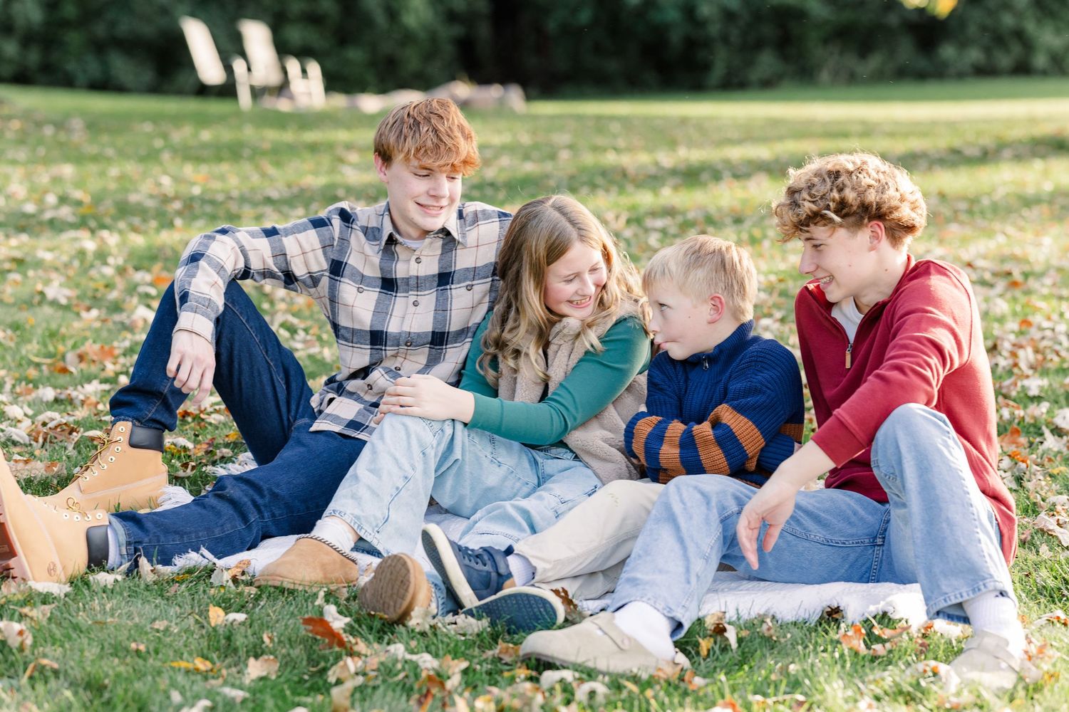 Four kids in plaid, red and striped fall clothes sit close together laughing on a leaf-strewn lawn in a sunny park.