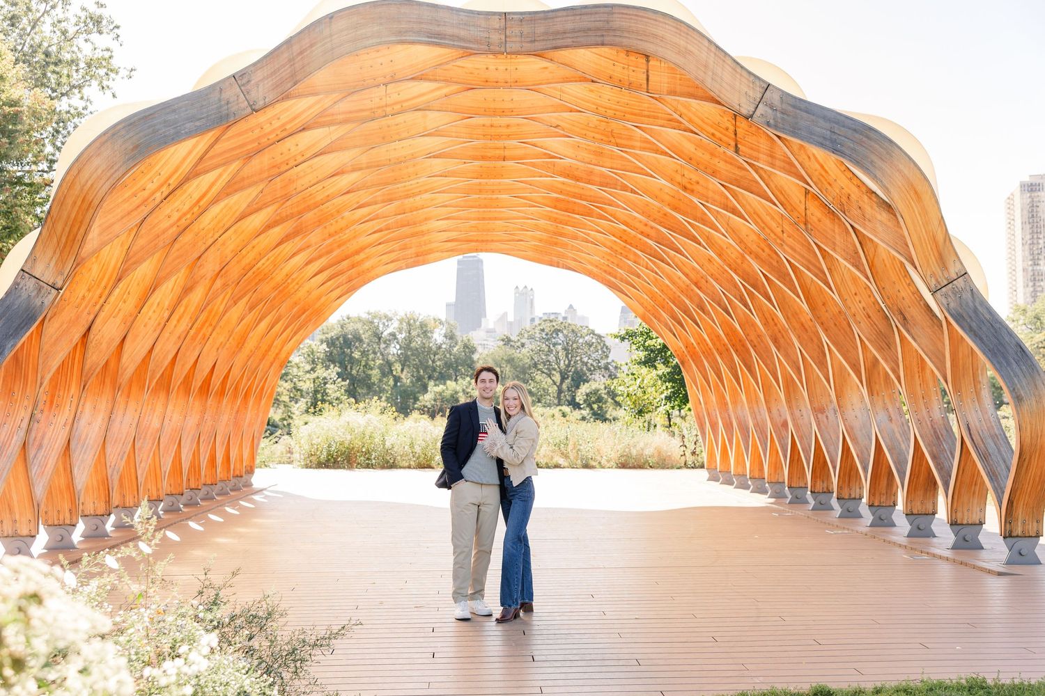 Couple embracing under a stunning orange wooden arch structure in a park with city skyline views.