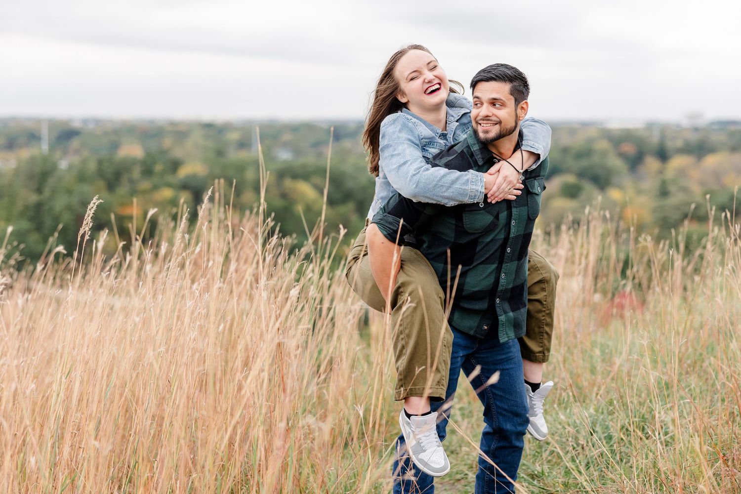 Laughing couple enjoying a playful piggyback ride through tall golden grass on a hilltop overlooking green trees on a cloudy autumn day.