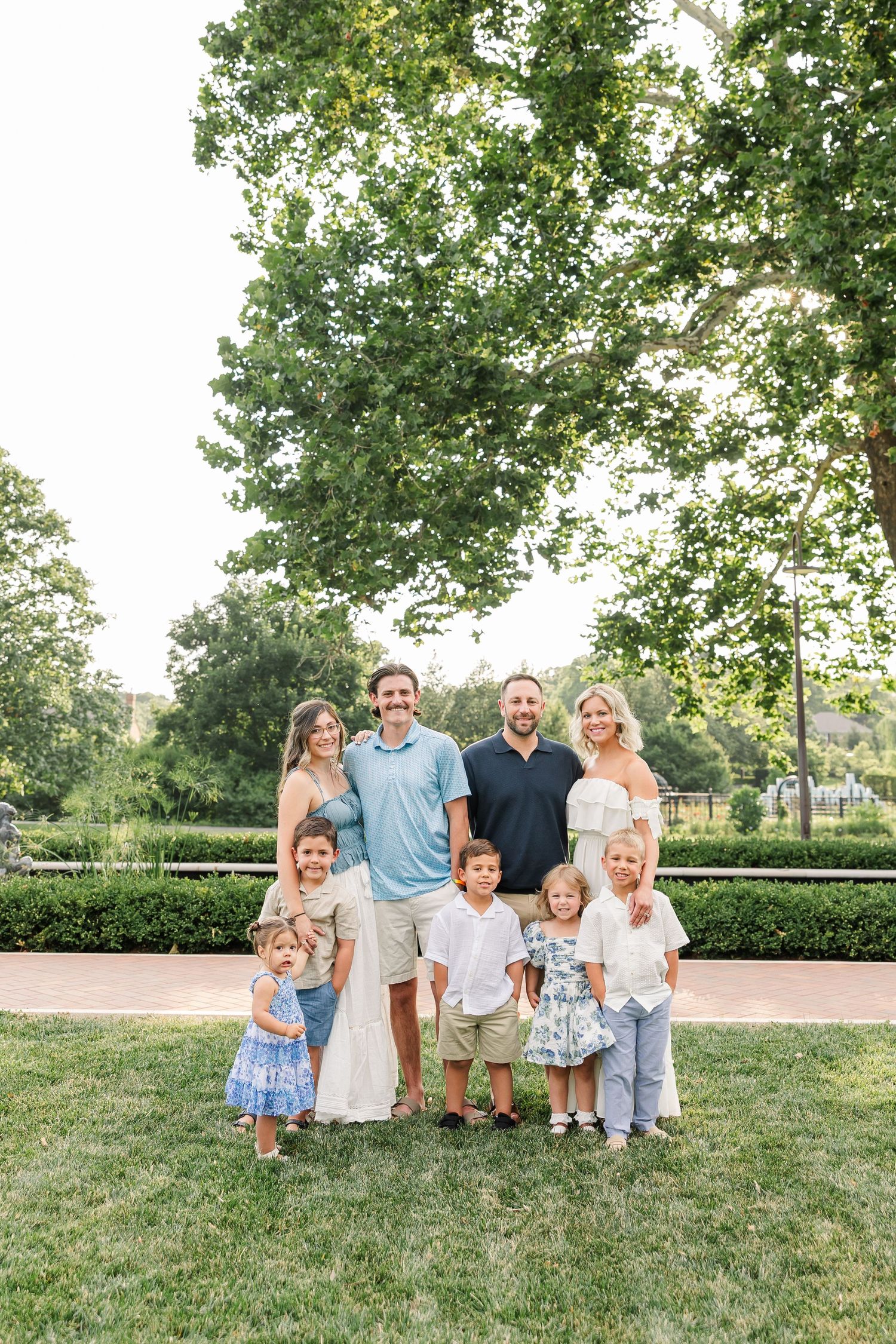 Extended family of adults and children posing together on green lawn with large trees in background.