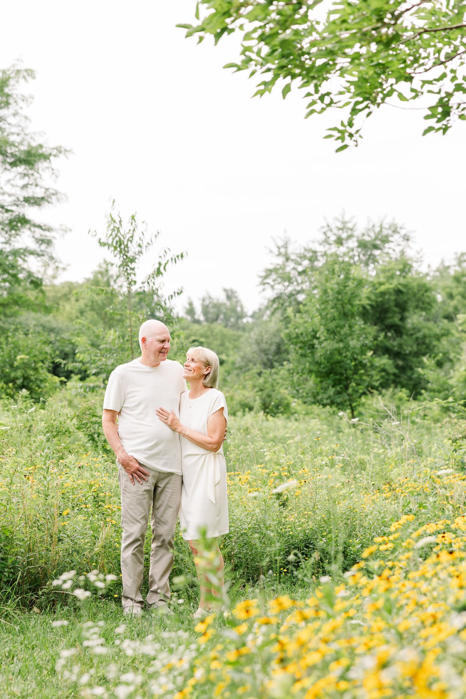 An elderly couple embraces in a lush green meadow filled with yellow wildflowers, sharing a loving glance outdoors.