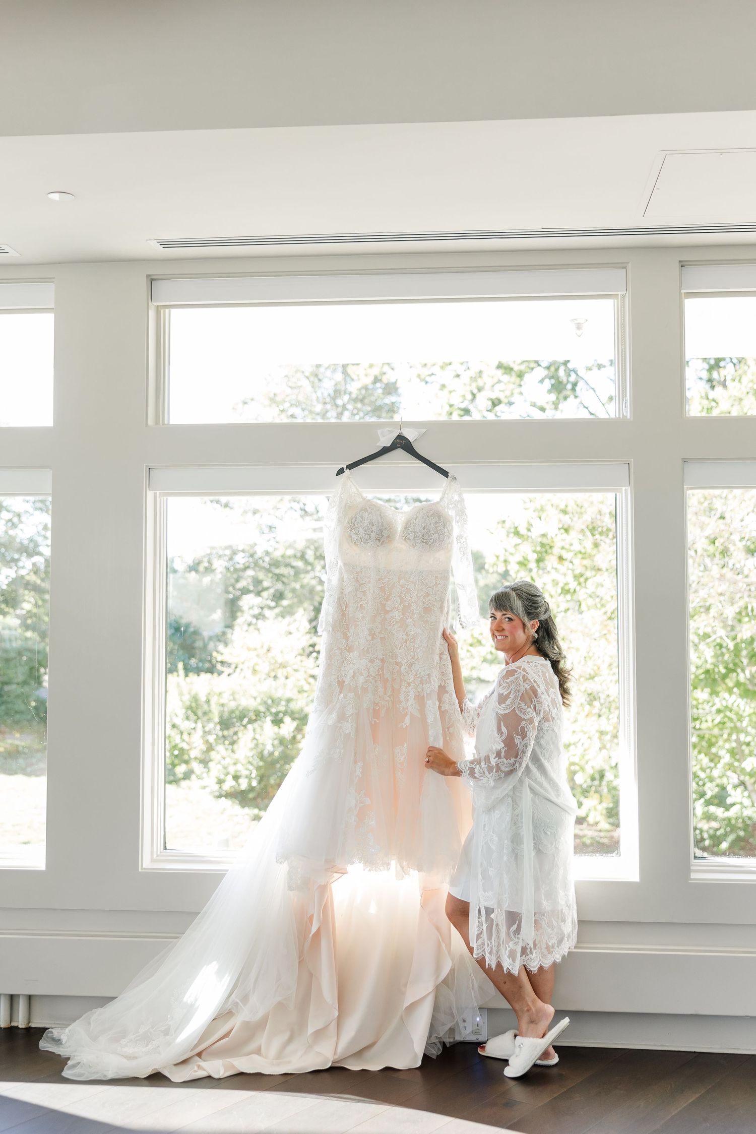 A bride in a white lace robe and slippers admires her lace wedding gown hanging by large sunlit windows.